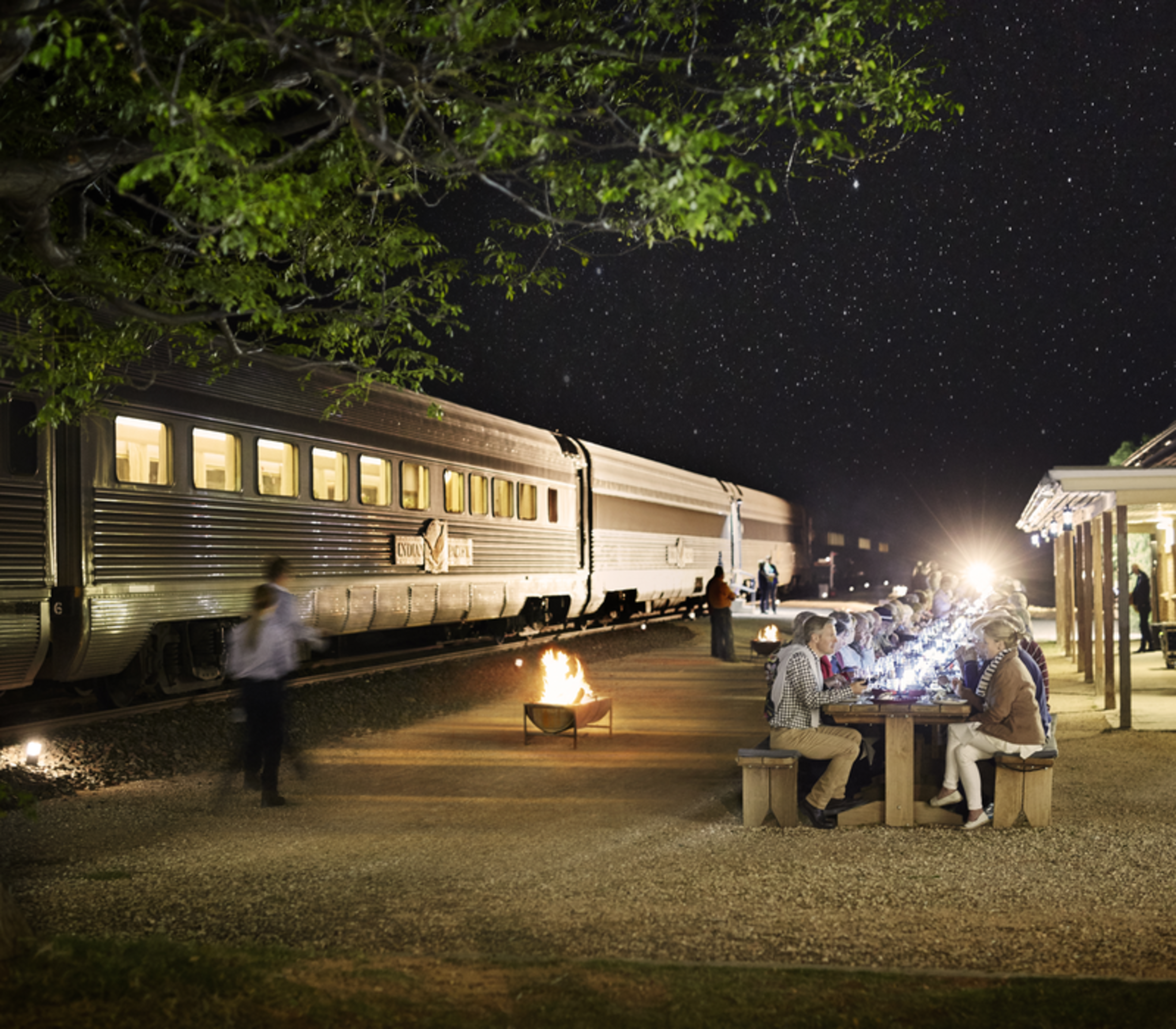 A silver Indian Pacific passenger train is parked alongside a remote station building at Rawlinna, where a large group of guests is seated at long outdoor wooden tables for a communal meal under a cloudy twilight sky.