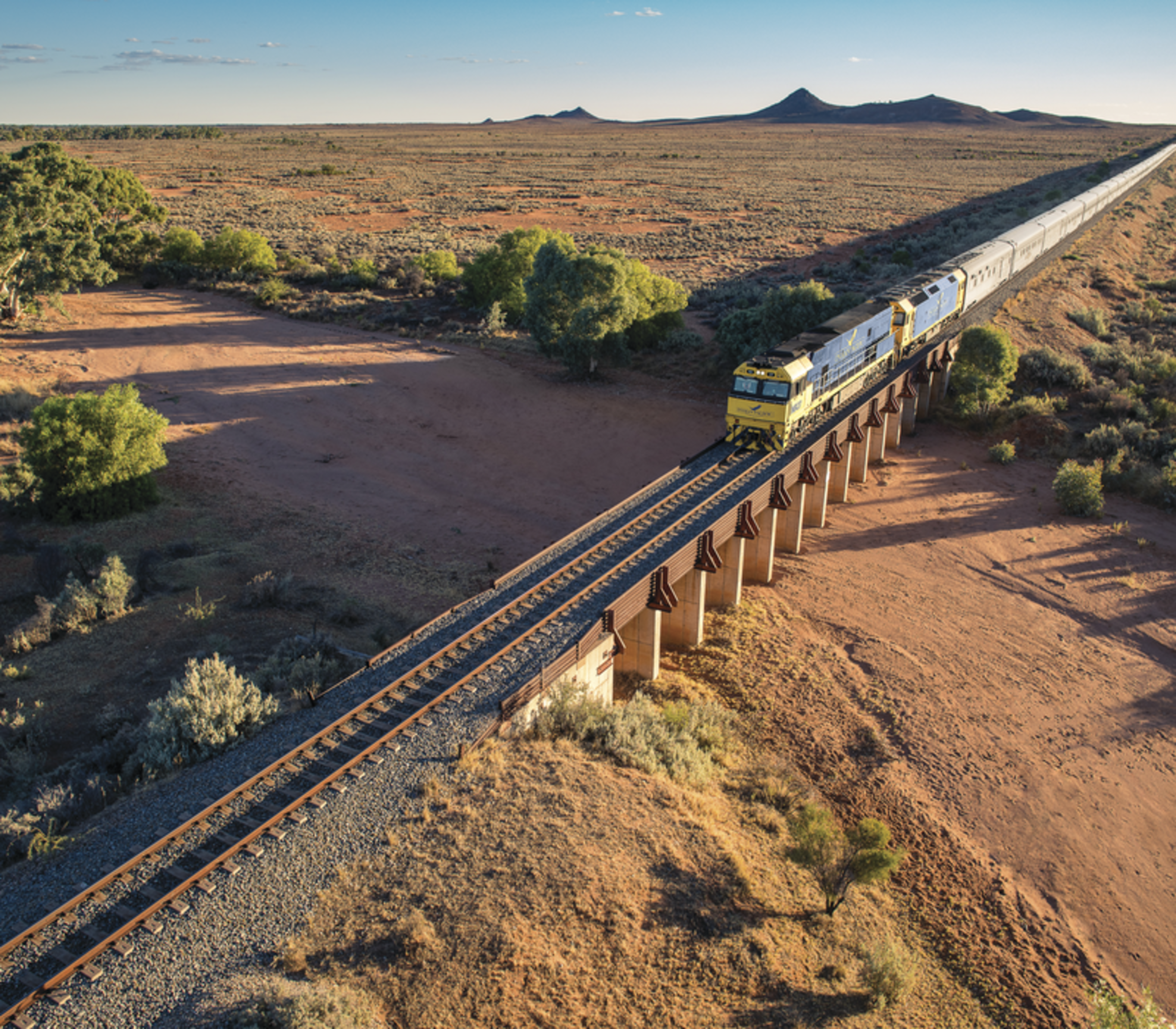 An aerial view of the long blue and yellow Indian Pacific passenger train winding along a curved track through vibrant, rolling emerald-green hills under a bright blue sky.