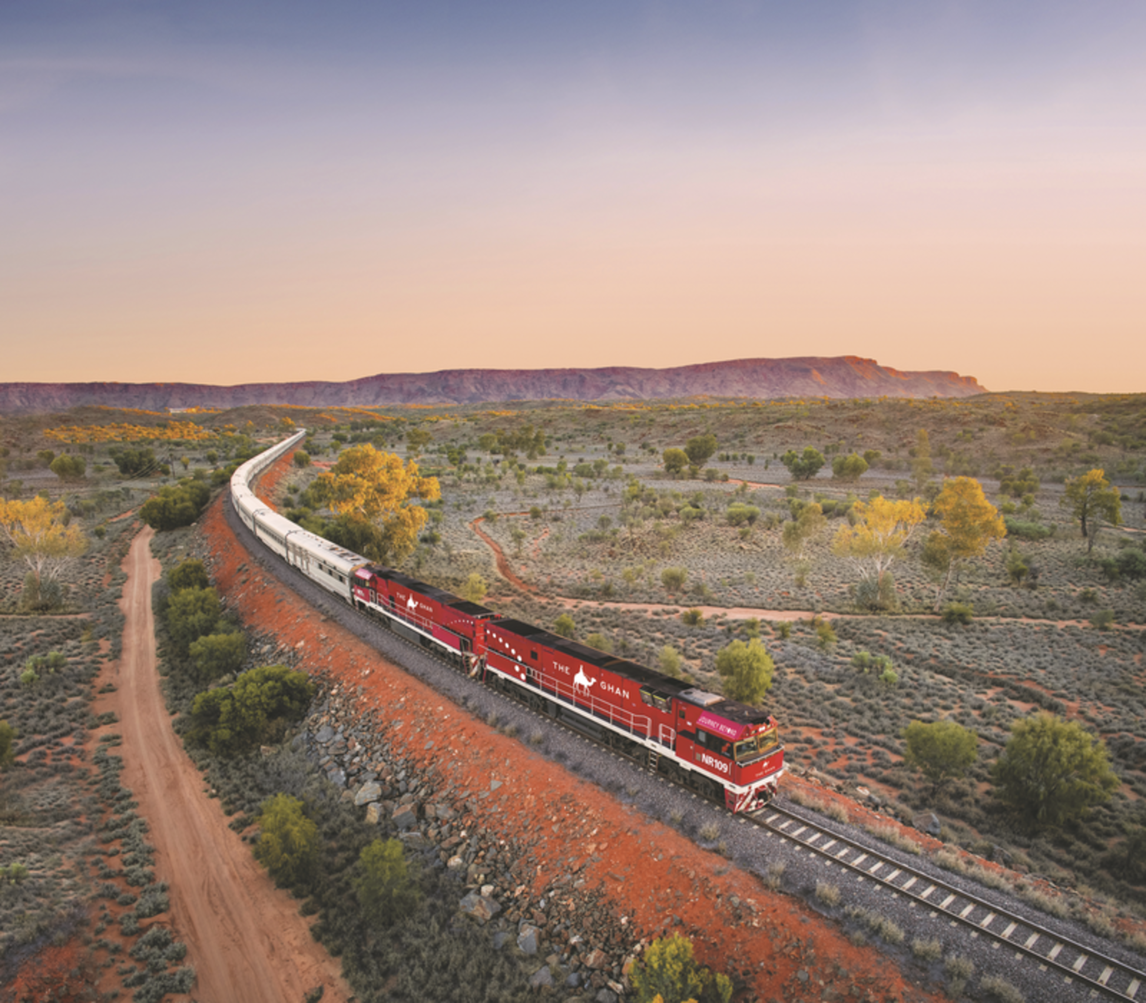High-angle view of the long Ghan train with its signature red engines curving through a vast desert landscape toward distant mountains.