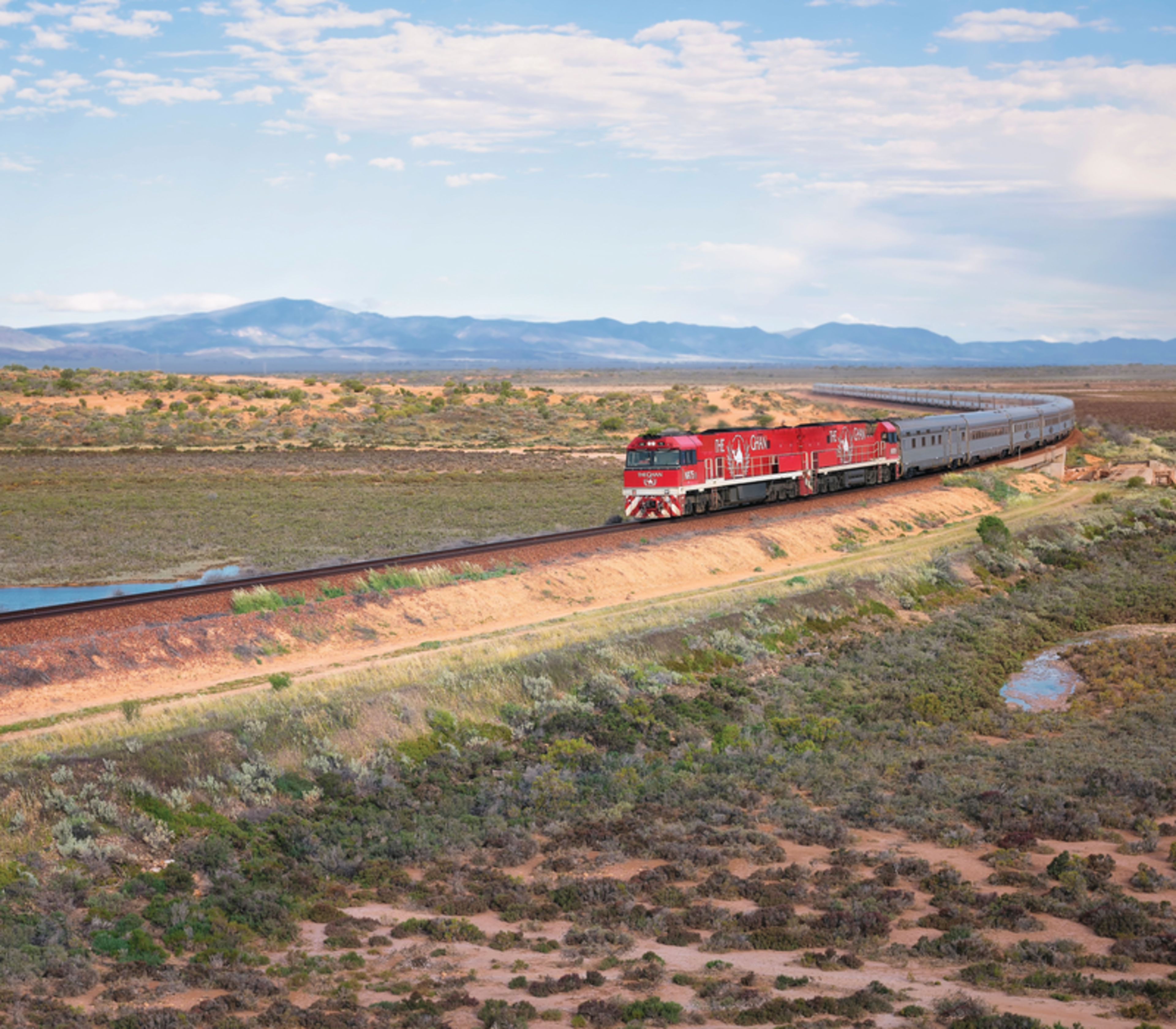 High-angle view of the long Ghan train with its signature red engines curving through a vast desert landscape toward distant mountains.
