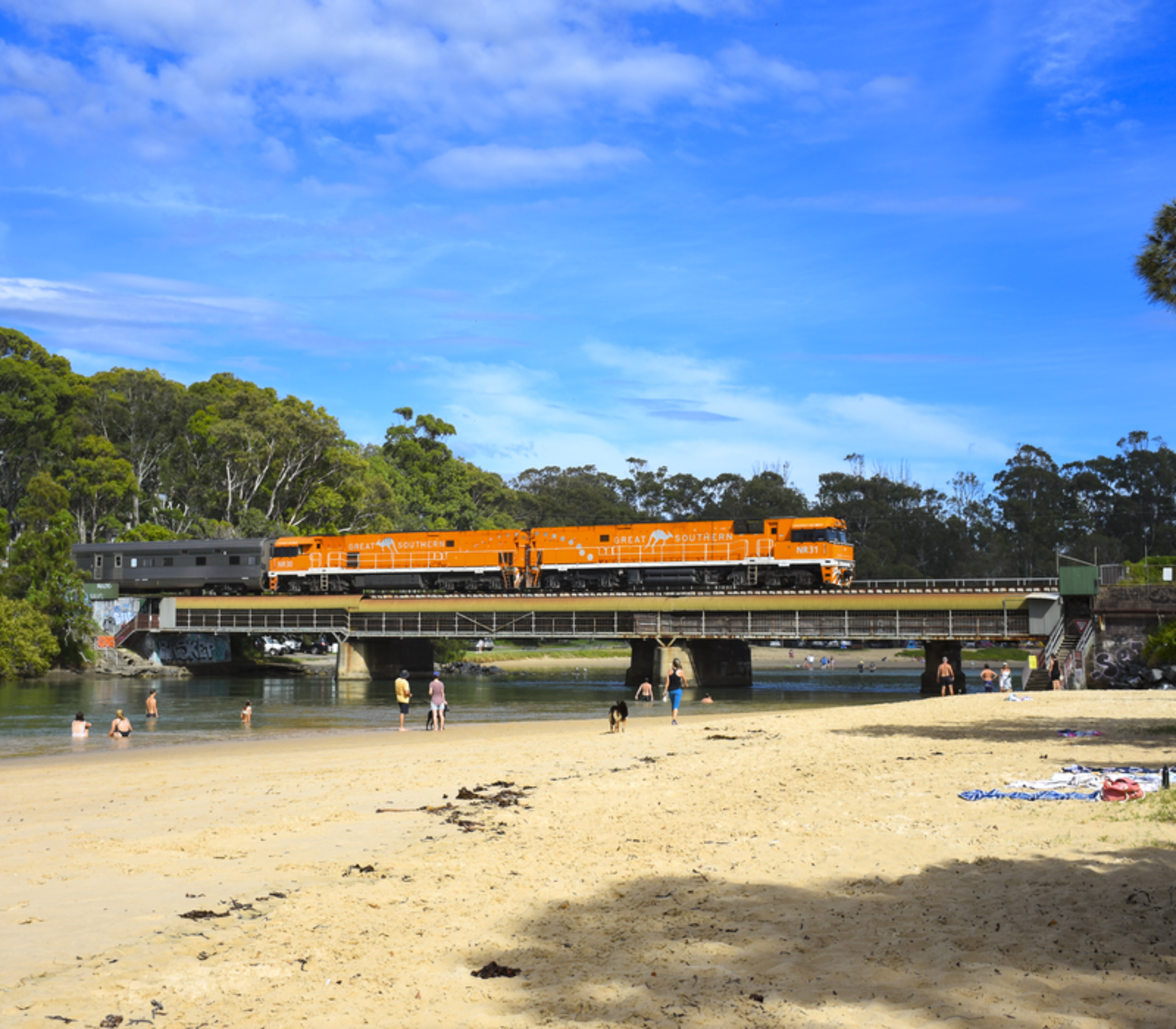 An orange passenger train crossing a bridge over a river with people on the beach in the foreground.