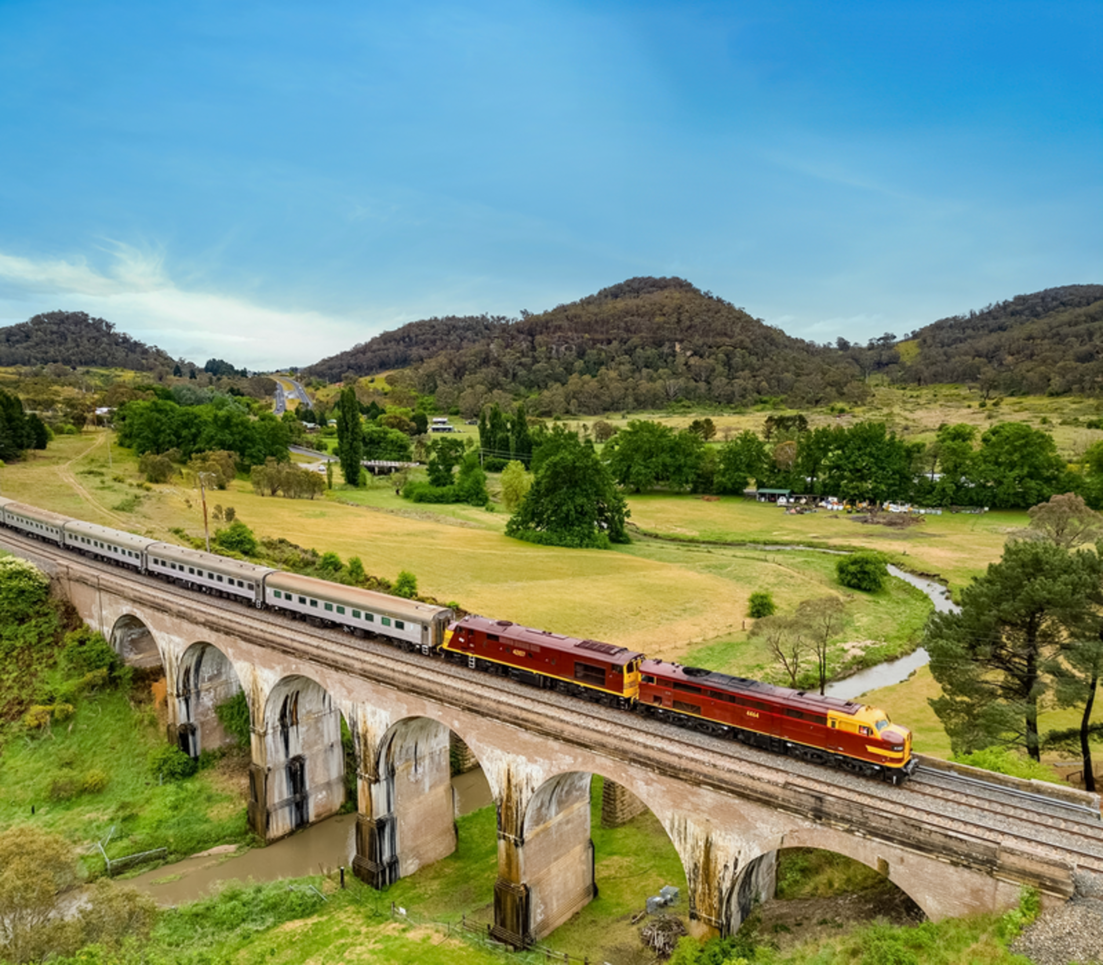 A heritage locomotive pulling a long silver train across a massive stone arch bridge in a valley.