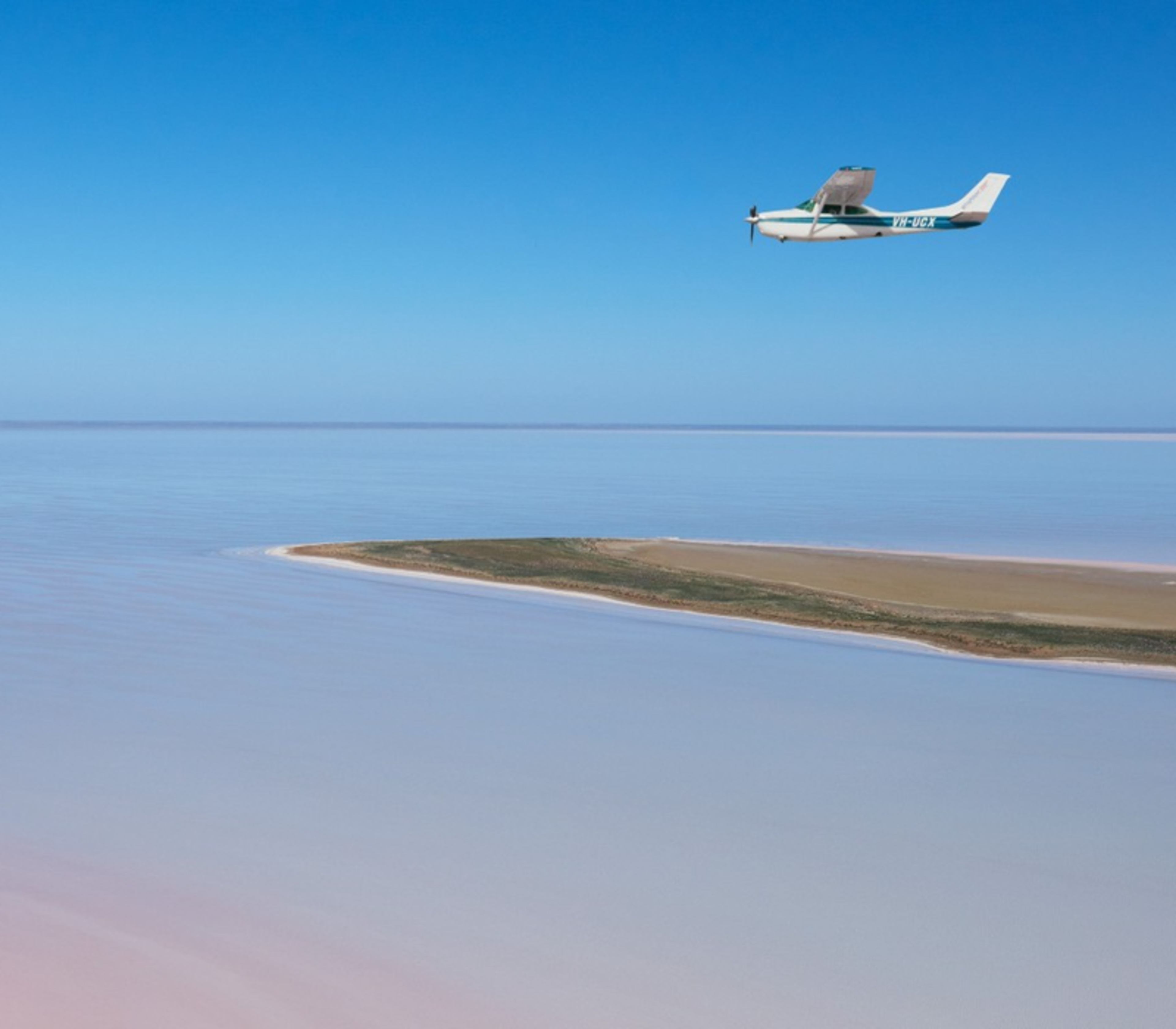 A small white scenic plane flying over the vast pink and white salt crust of Lake Eyre.
