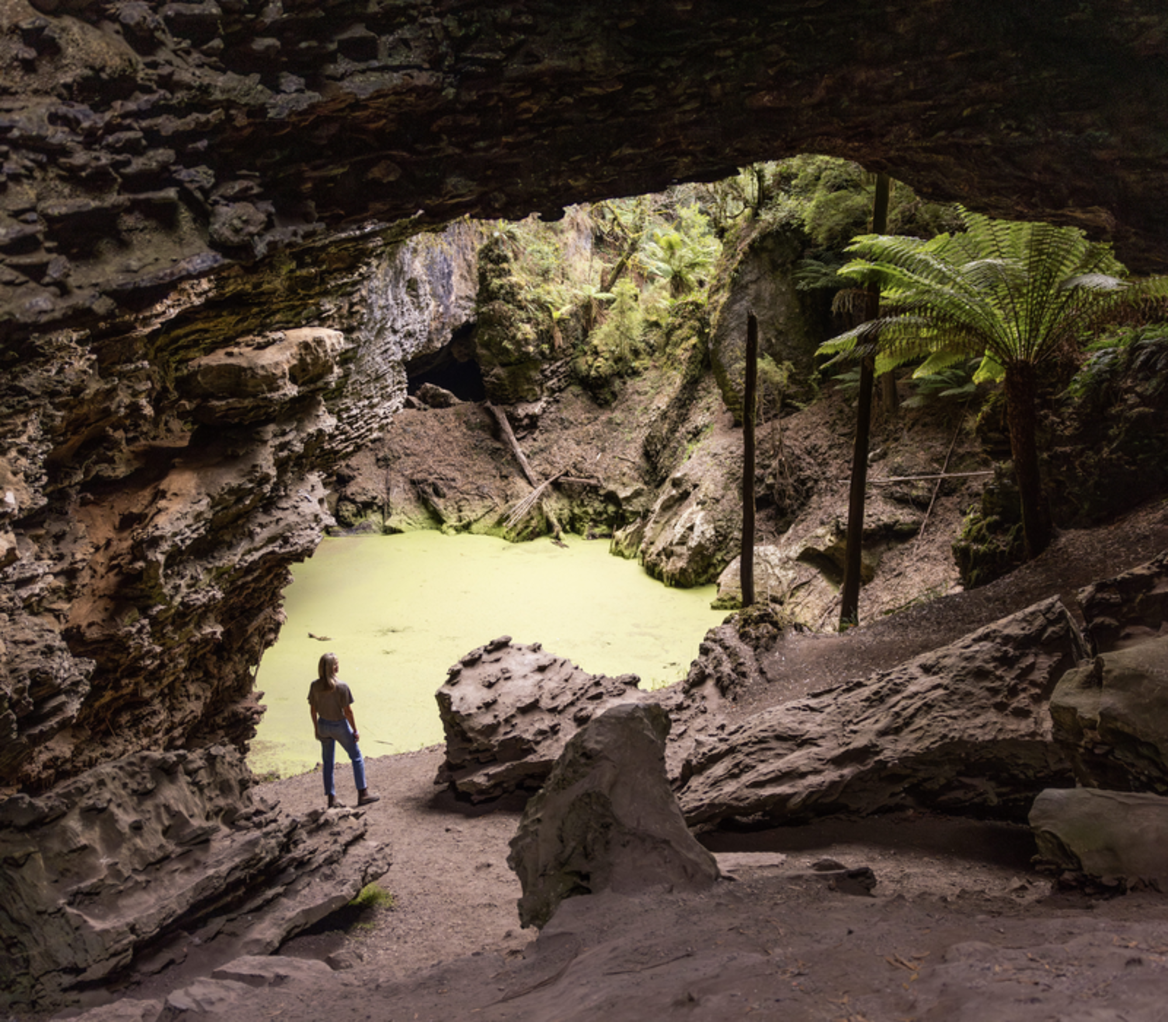 A person looking at a natural rock arch over a lime-green pond surrounded by rainforest.