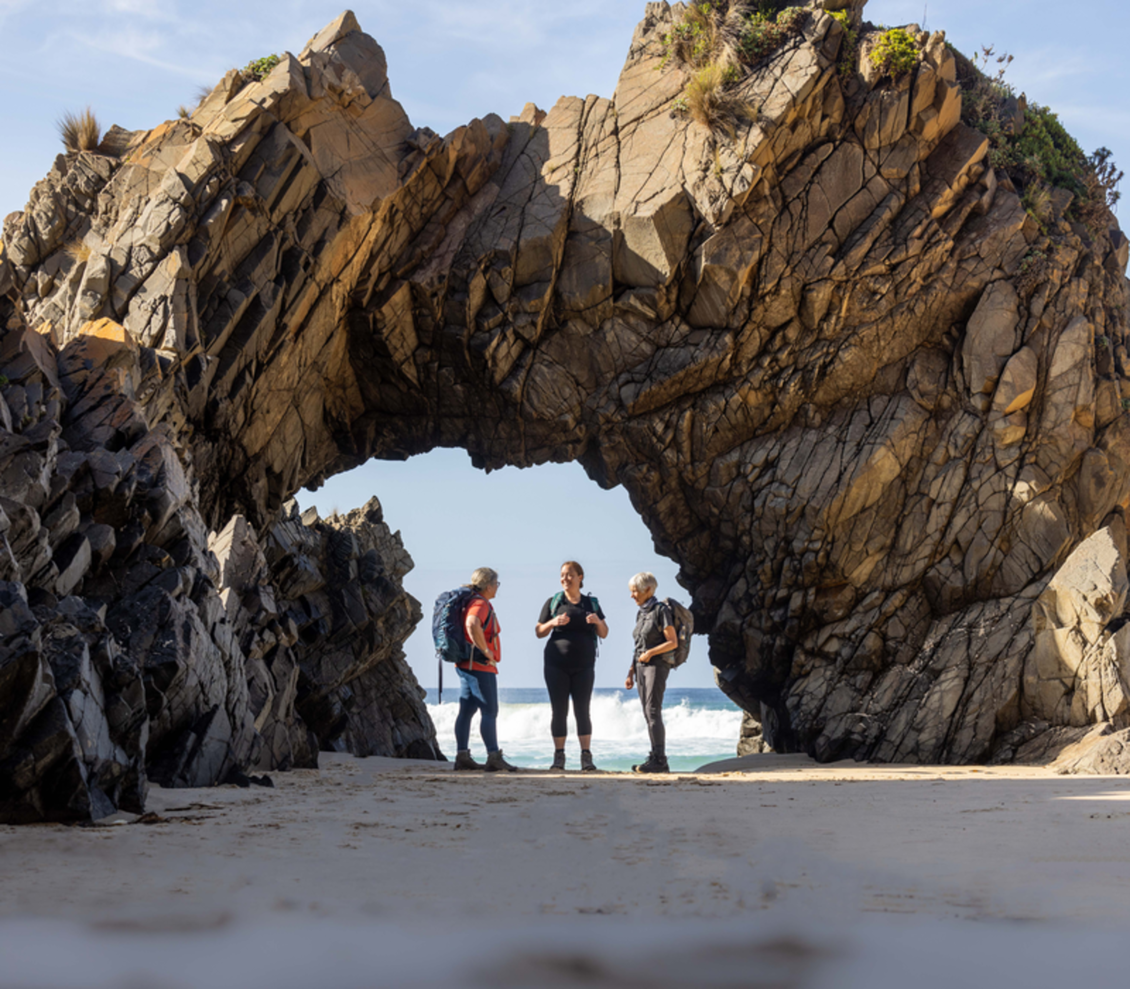 Travelers standing under a large natural rock formation arch on a beach in Tasmania.