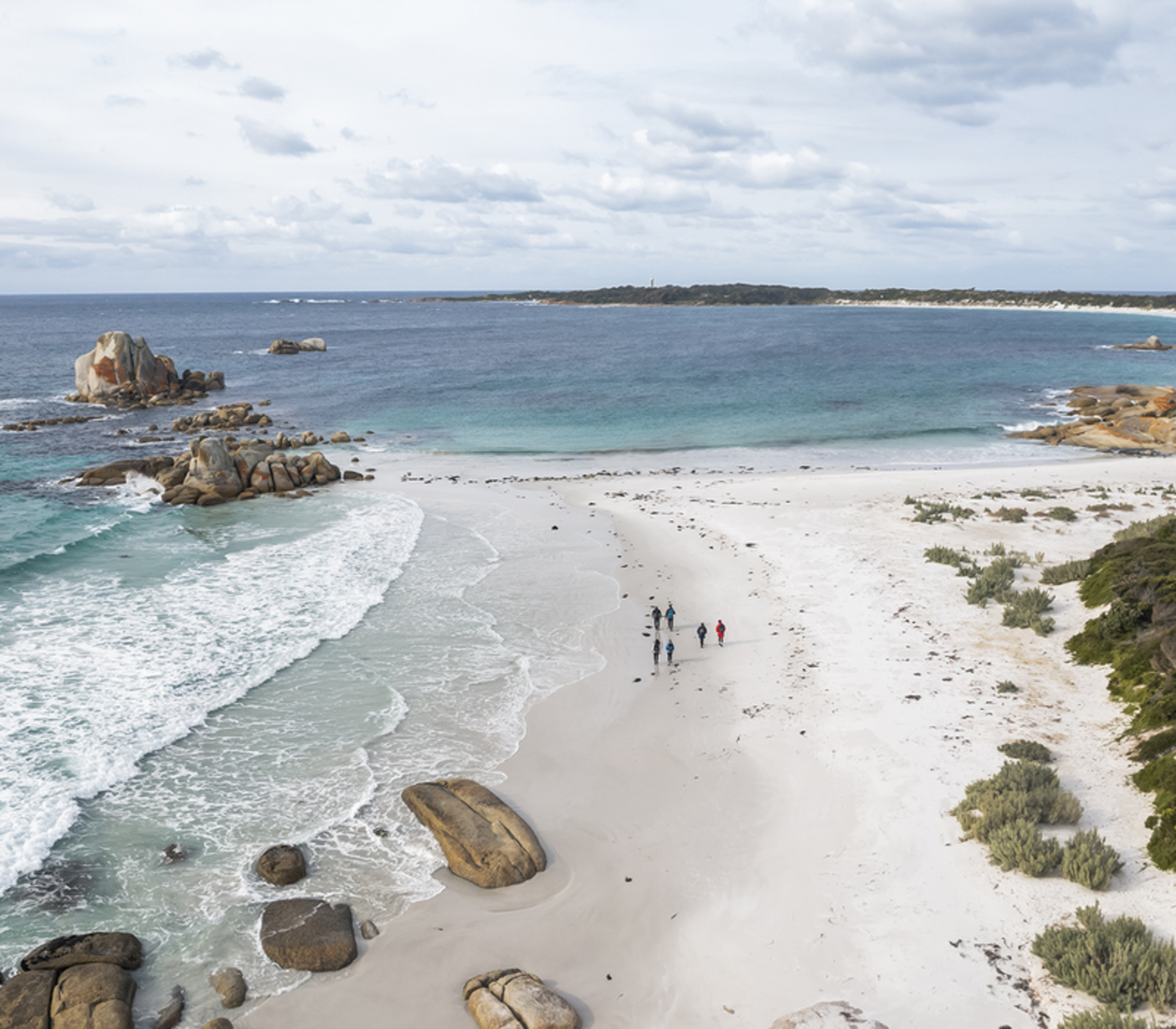 Aerial view of a small group of people walking along a white sand beach next to turquoise ocean and orange boulders.
