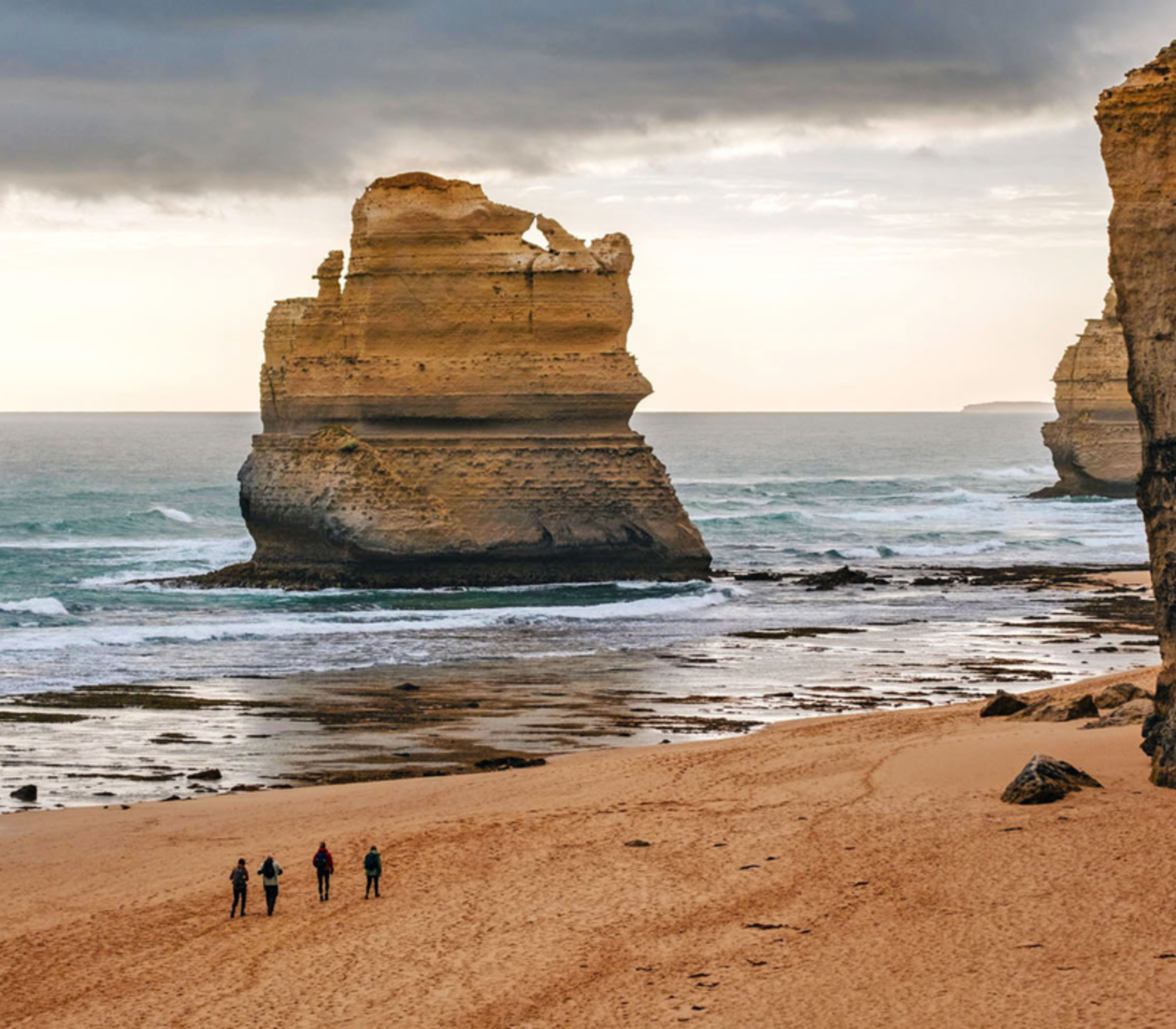 A small group of hikers walking on a wide sandy beach next to massive limestone cliffs.