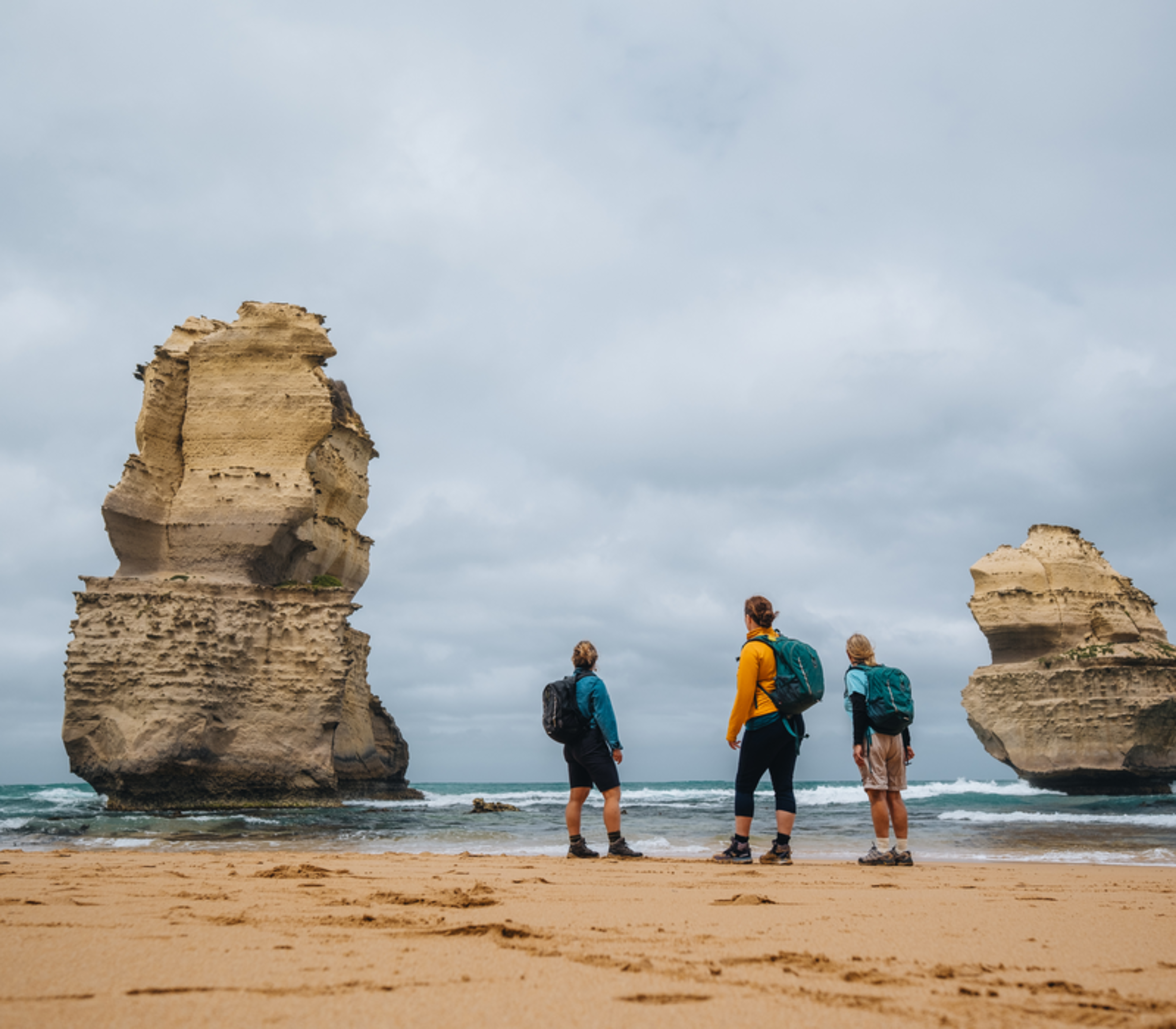 Three people standing on a sandy beach looking up at massive limestone stacks in the sea.