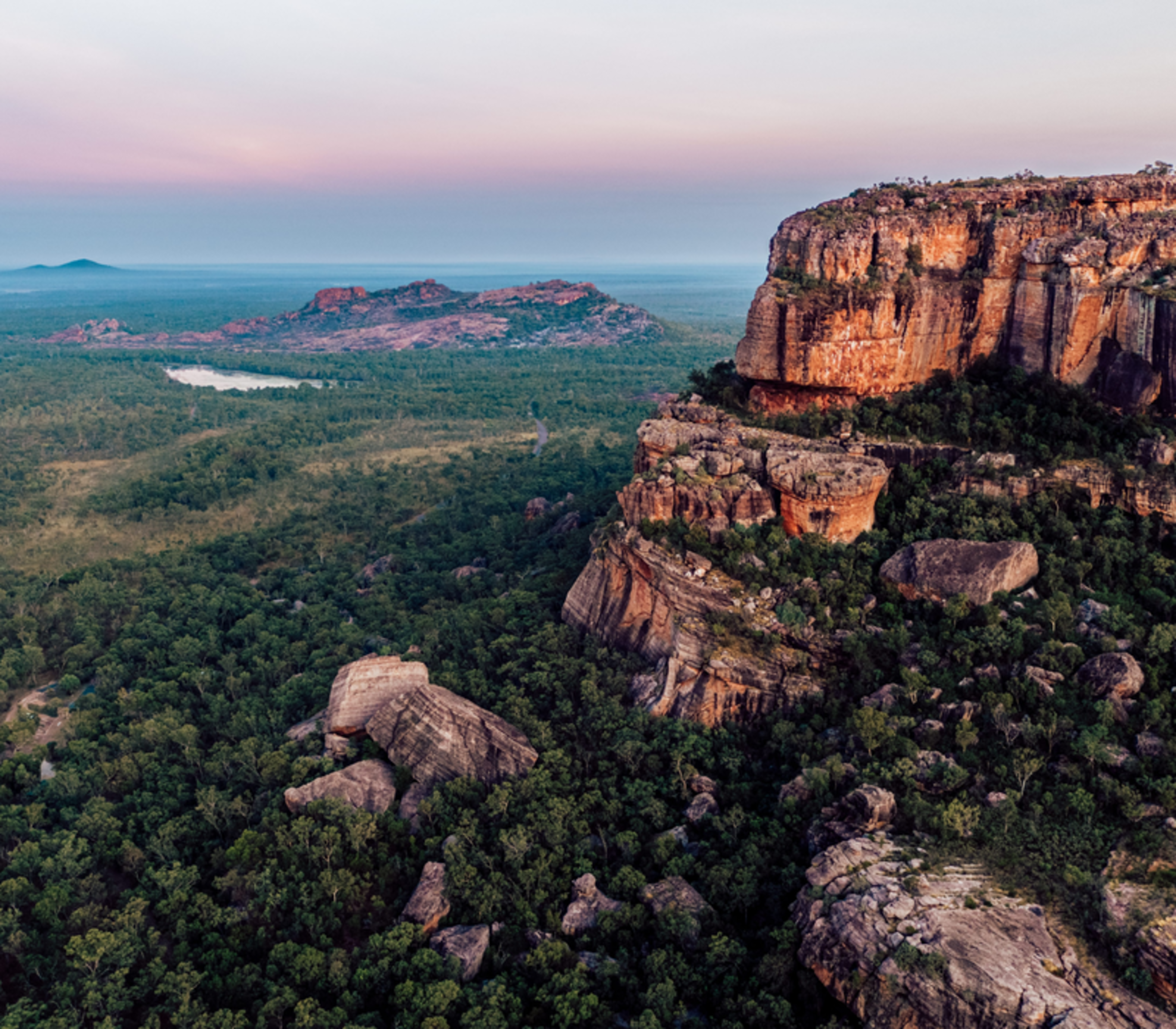 A breathtaking aerial shot of the rugged red sandstone cliffs of Burrungkuy (Nourlangie) at dusk, with the vast, green savanna woodlands of Kakadu National Park stretching toward the horizon under a soft pink and purple sky.