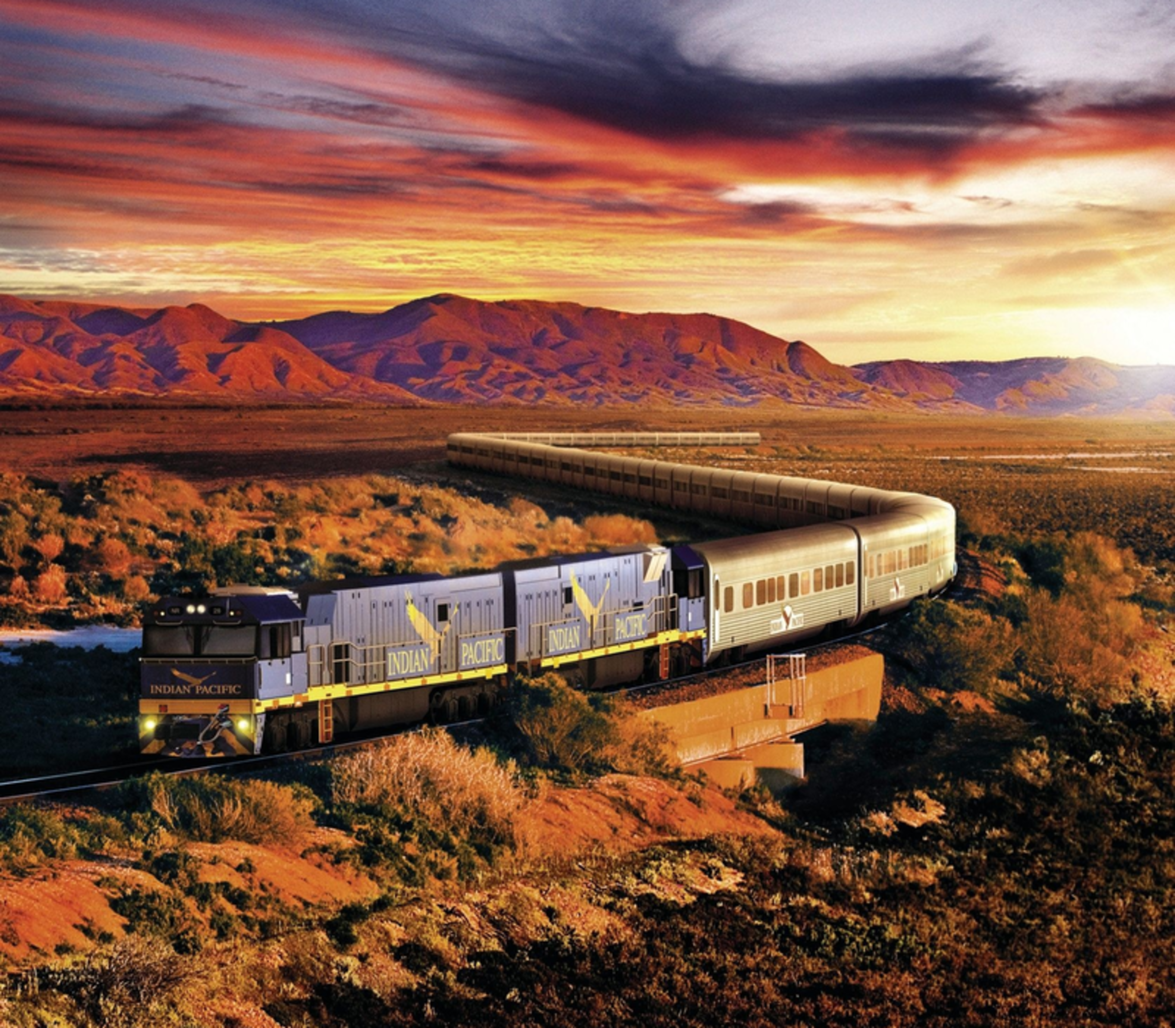 The Indian Pacific passenger train curves through the arid outback of South Australia under a dramatic orange and purple sunset sky.