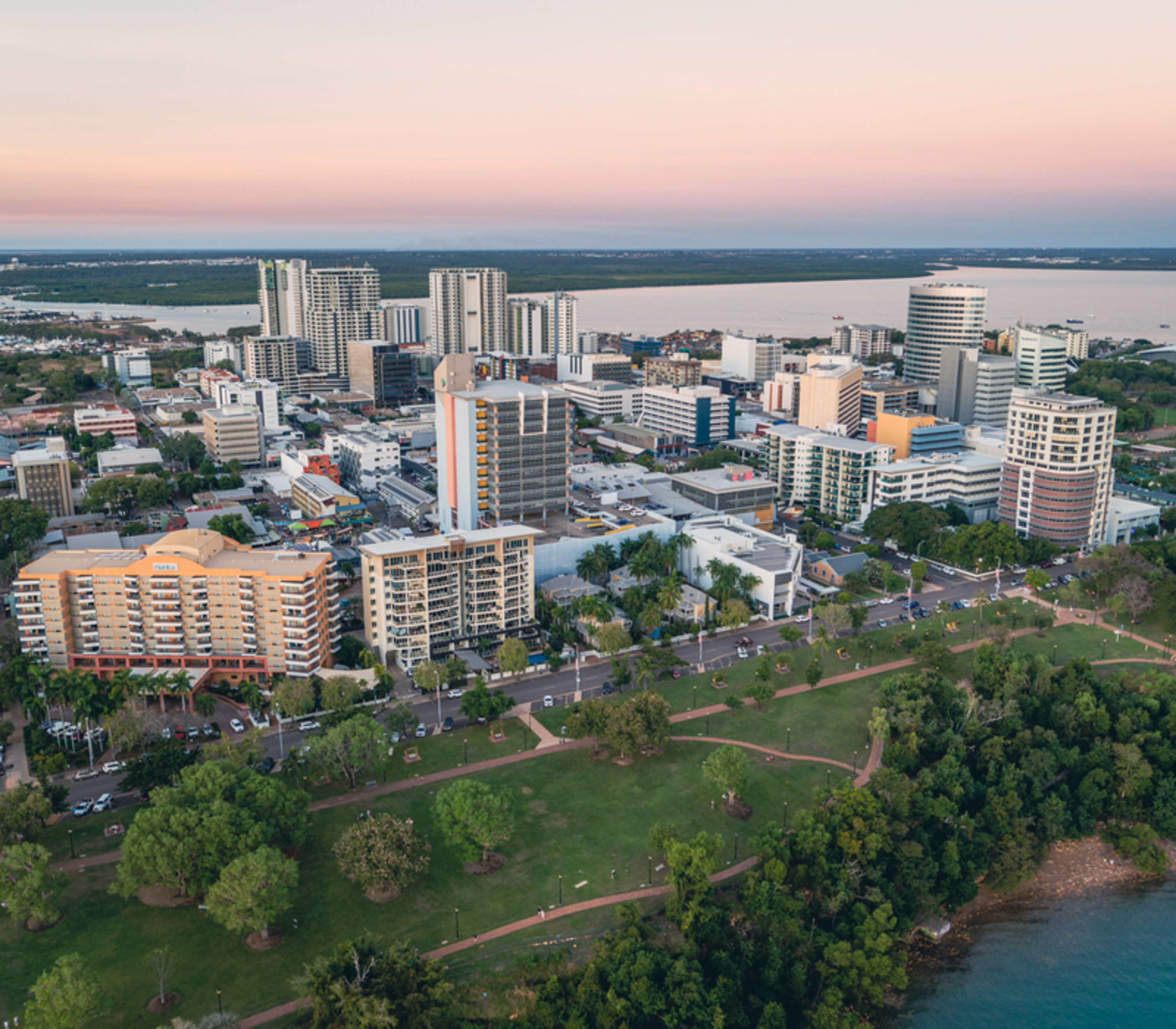 An expansive aerial view of the Darwin city skyline and lush green Esplanade parklands at dusk, overlooking the Timor Sea.