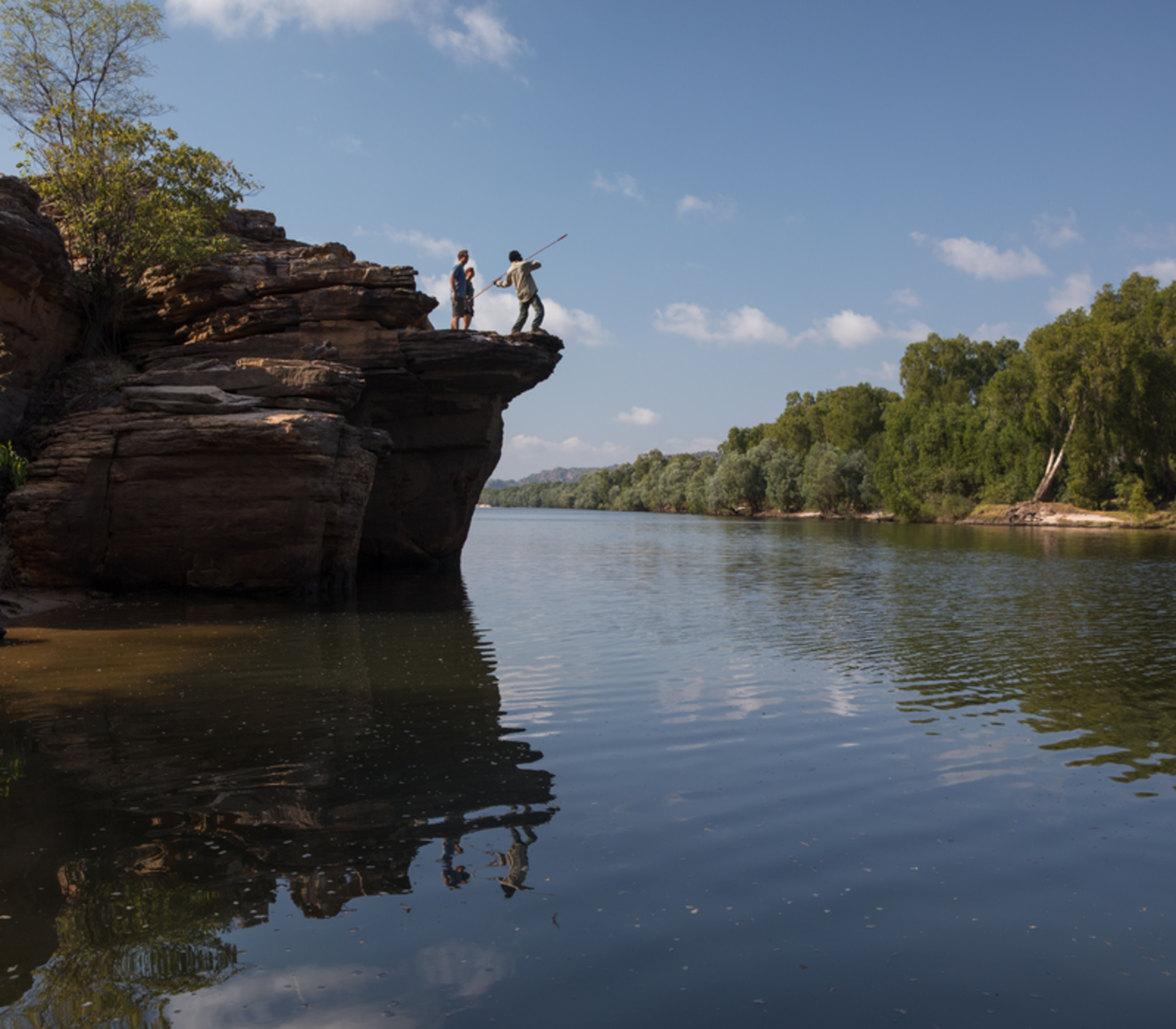 An Aboriginal guide stands on a dramatic sandstone ledge above the calm East Alligator River, holding a traditional spear and demonstrating its use to a man and woman standing nearby in Kakadu.