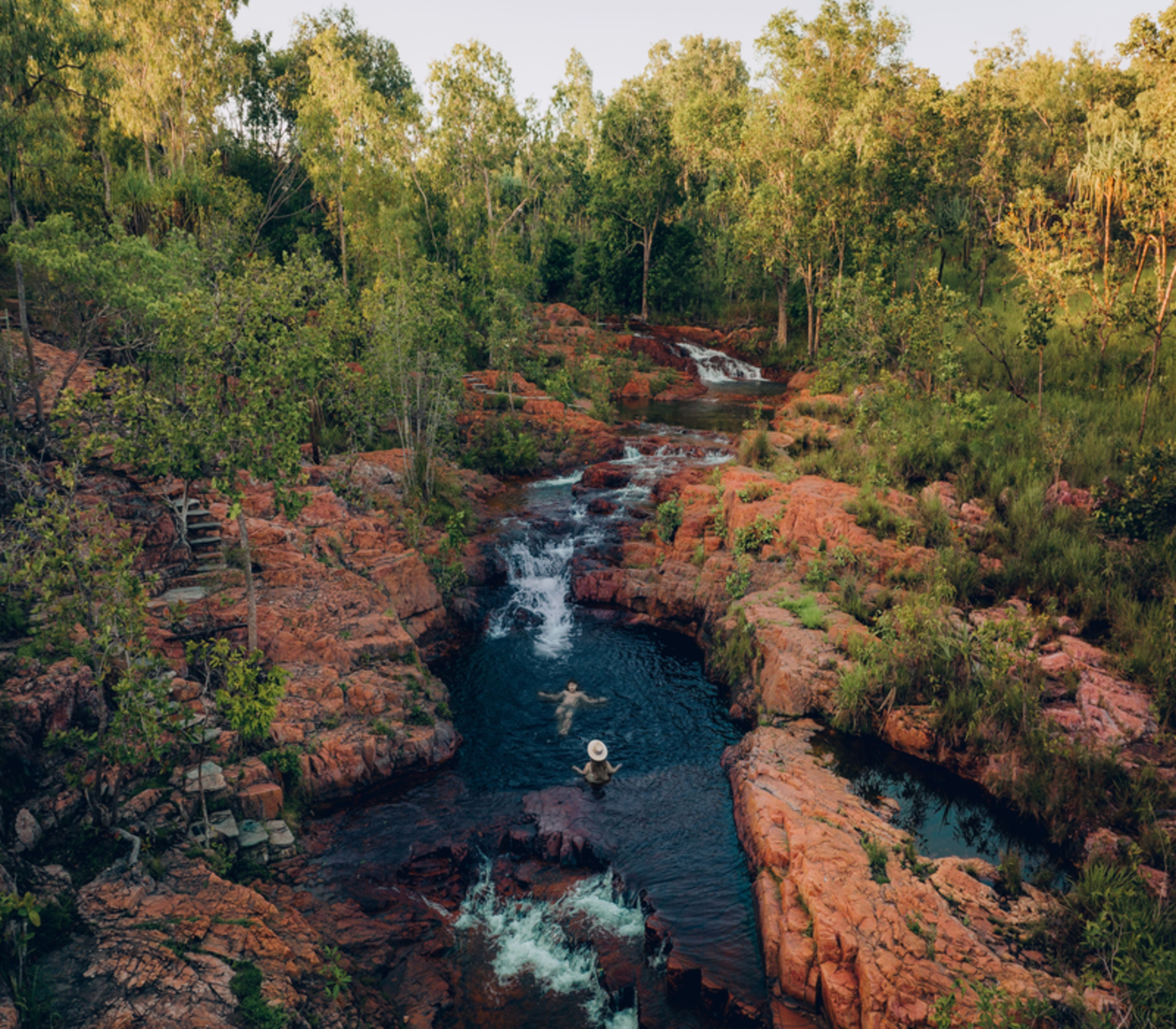 People swimming in tiered natural waterfalls and rock pools surrounded by green forest in Litchfield National Park.