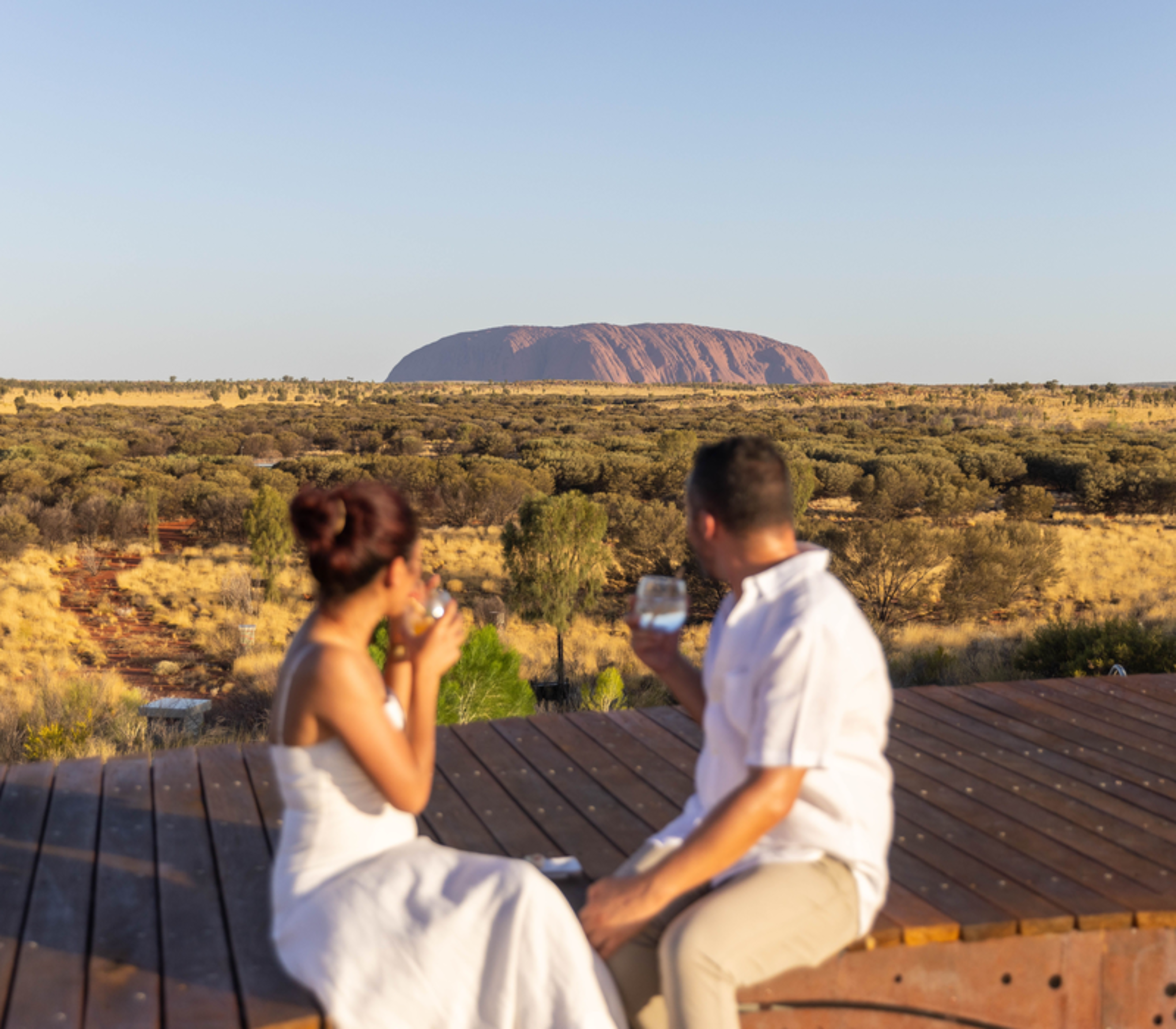 A man and woman sit on a wooden deck in the foreground, blurred, while holding drinks and looking out over the vast desert toward the iconic red monolith of Uluru under a clear sky.