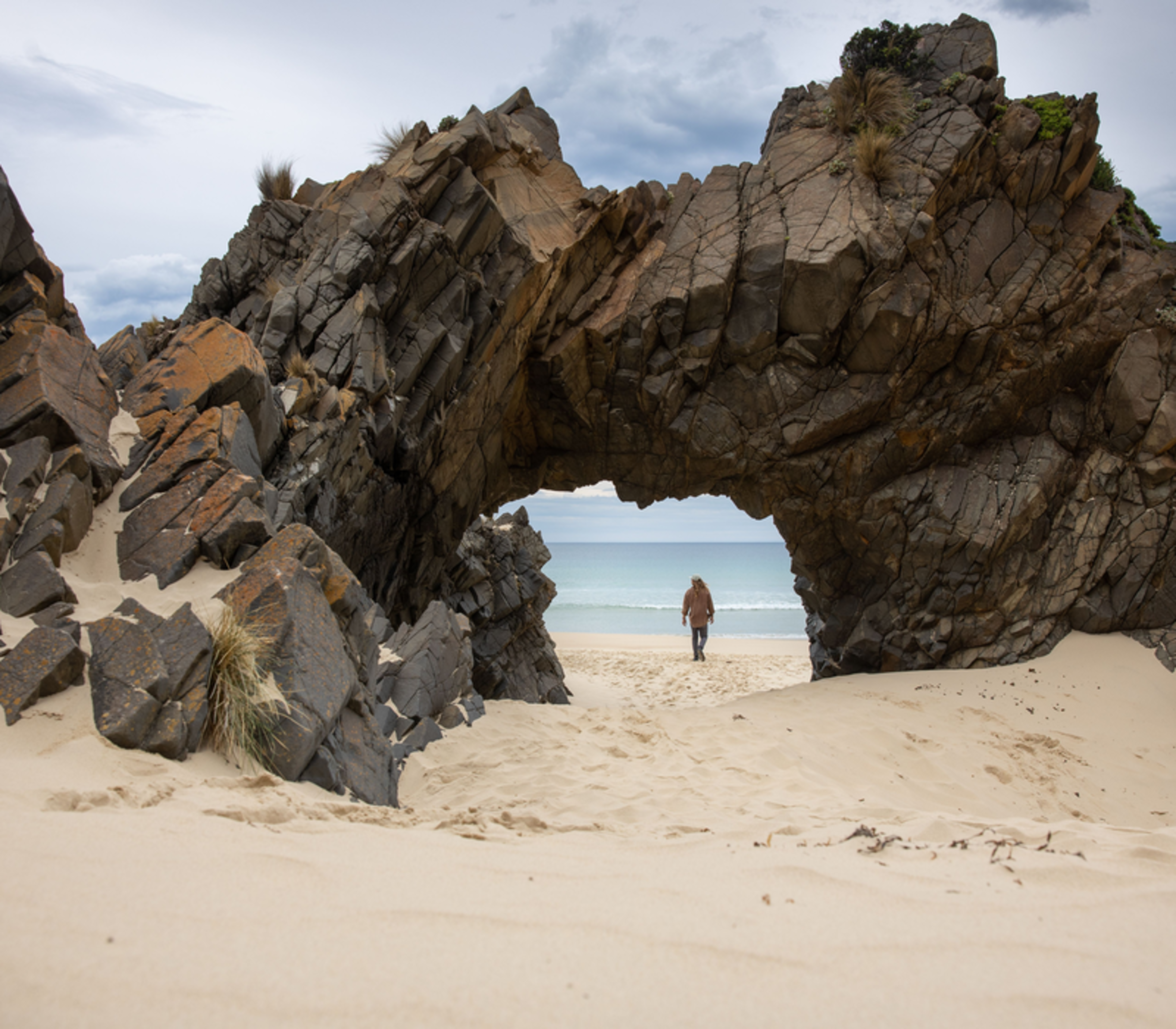 A natural rocky archway stands on a sandy beach at Mars Bluff, with a hiking trail winding through coastal heath and views of the blue ocean in the background.