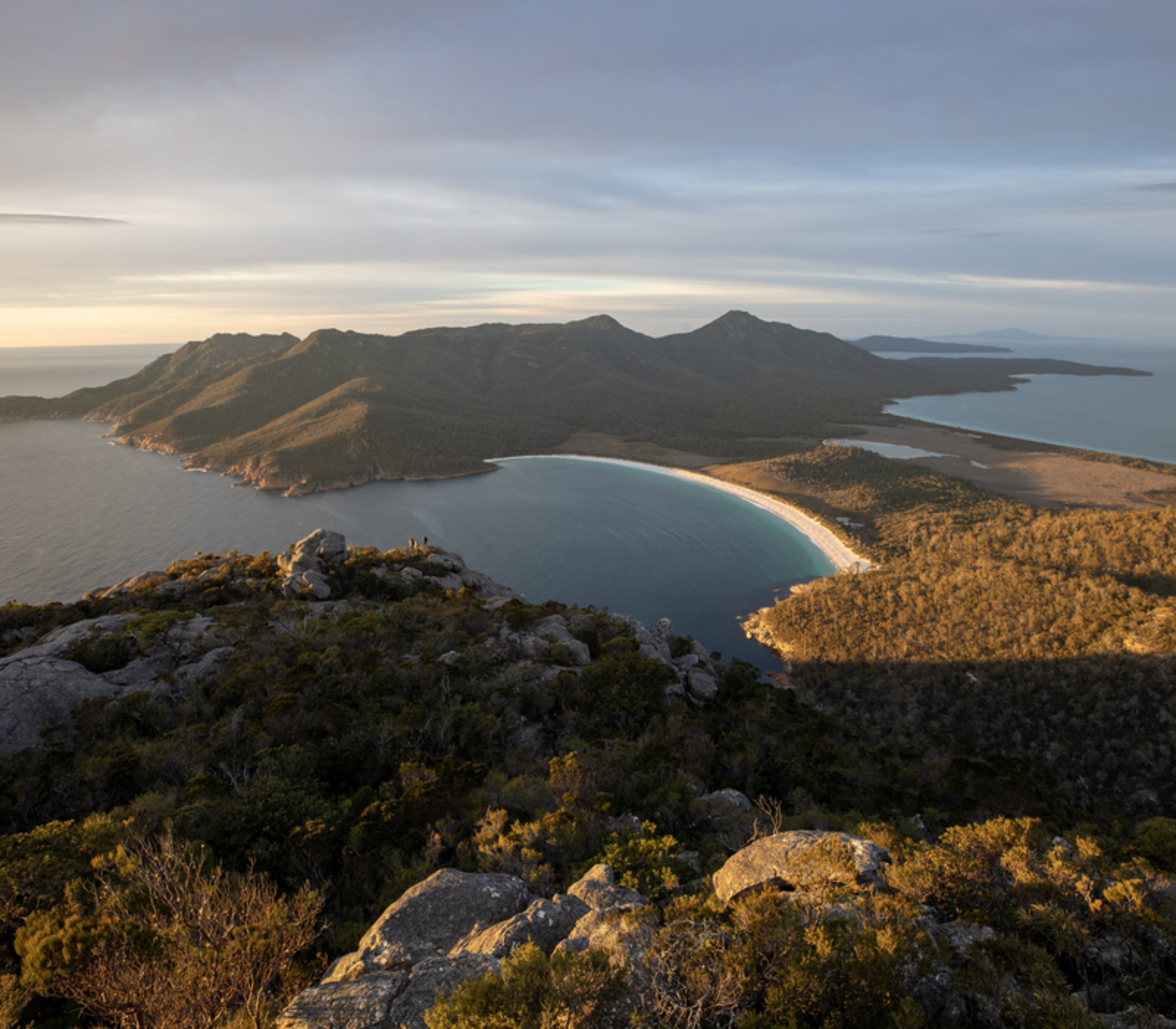 Panoramic high-angle view of a crescent-shaped white sand beach and turquoise bay surrounded by forested mountains under a soft sunrise sky.