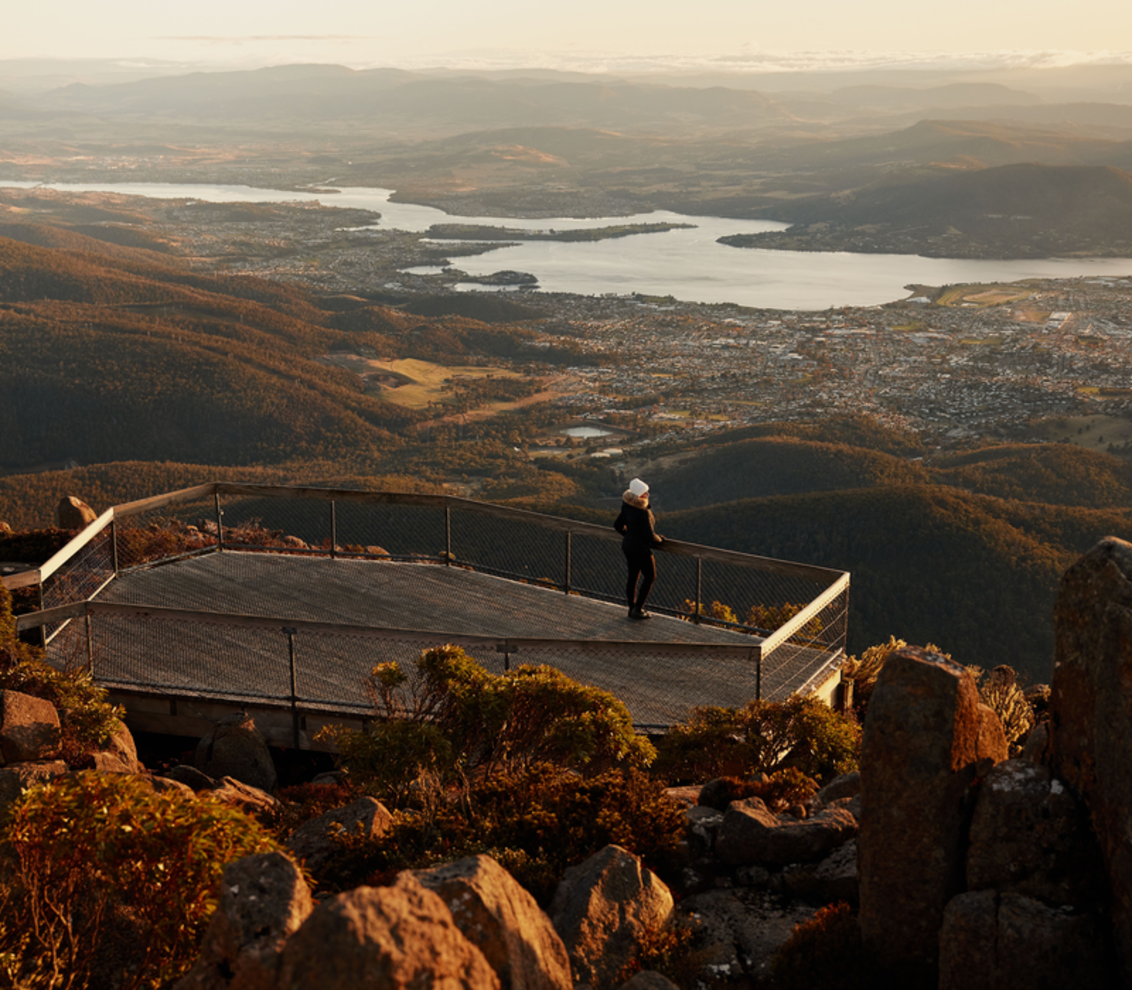 Traveler at the Mount Wellington summit observation deck during golden hour overlooking the Hobart region landscape.