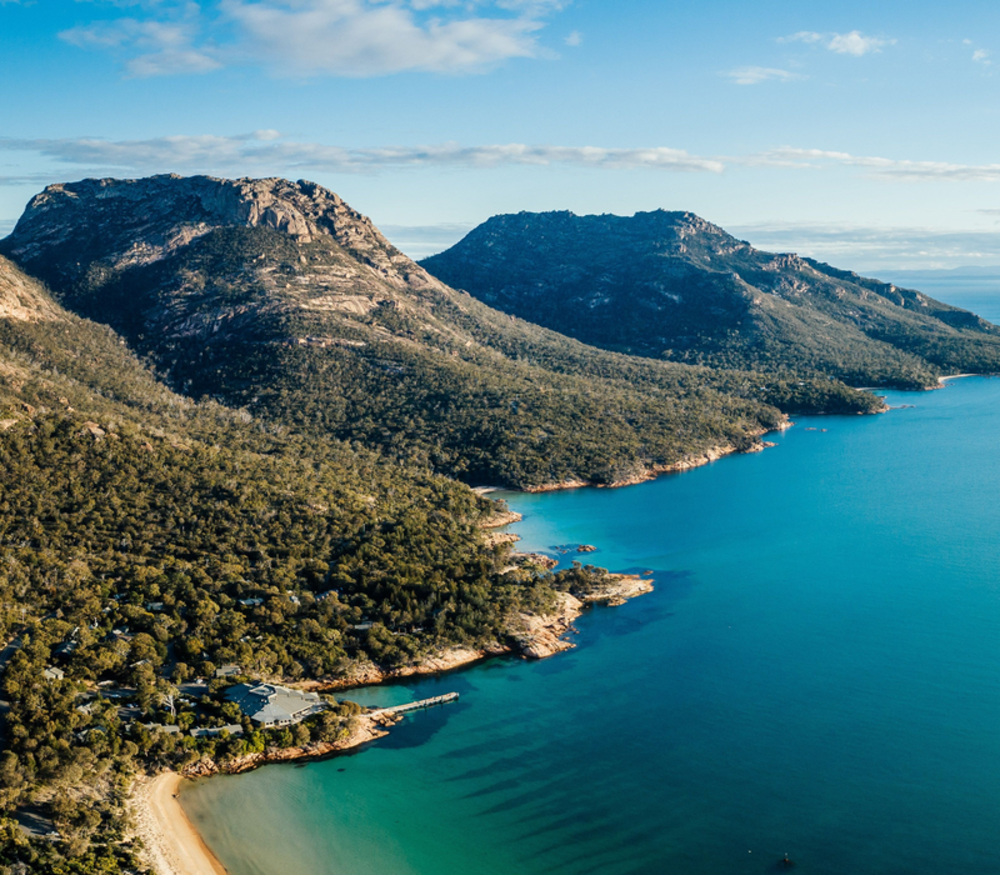 erial panorama of the Freycinet Peninsula coastline, featuring the main complex of Freycinet Lodge tucked into the dense green forest at the water's edge of Great Oyster Bay, with the striking pink granite peaks of the Hazards mountain range rising in the