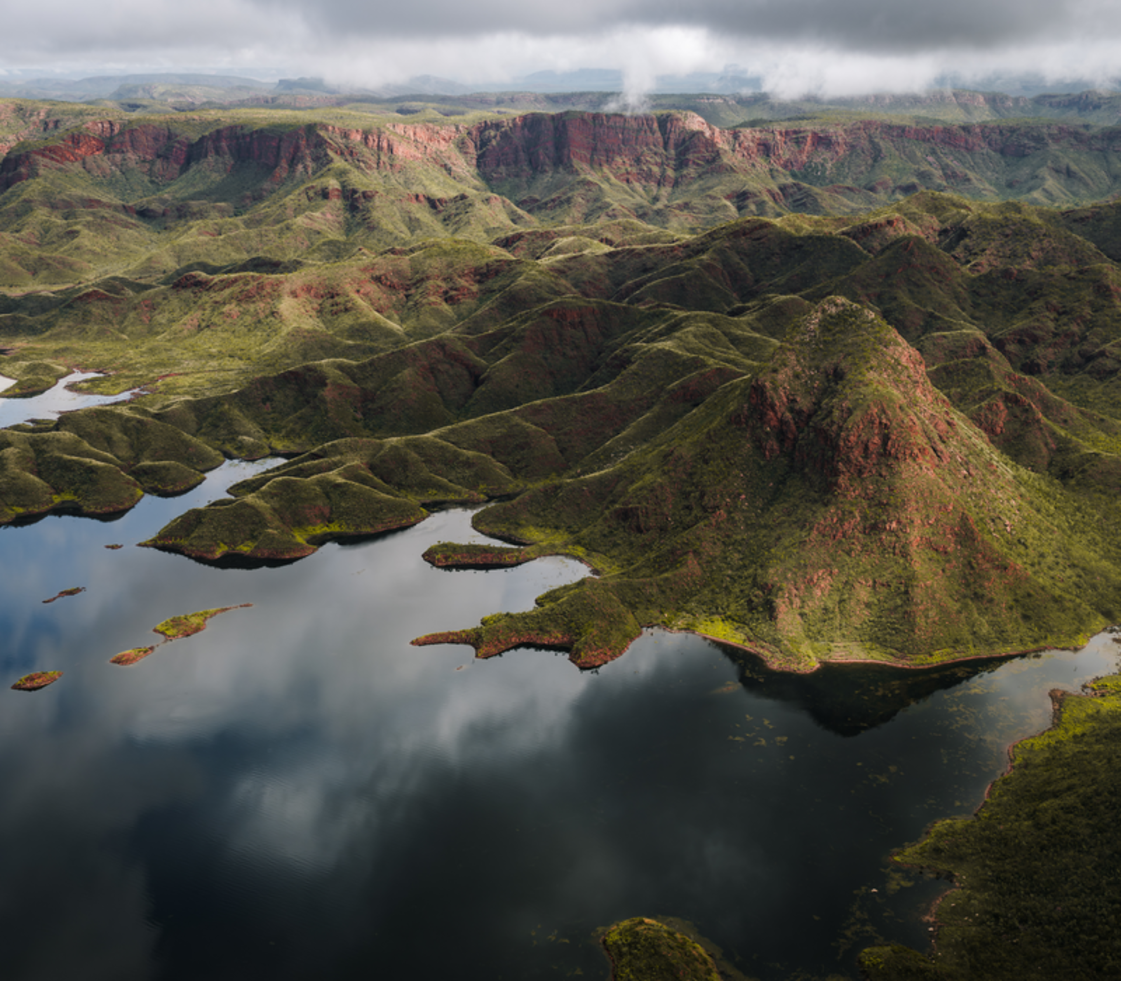 A high-angle aerial view of a green and red hilly landscape meeting a lake, with low-hanging white clouds and shadows across the hills.