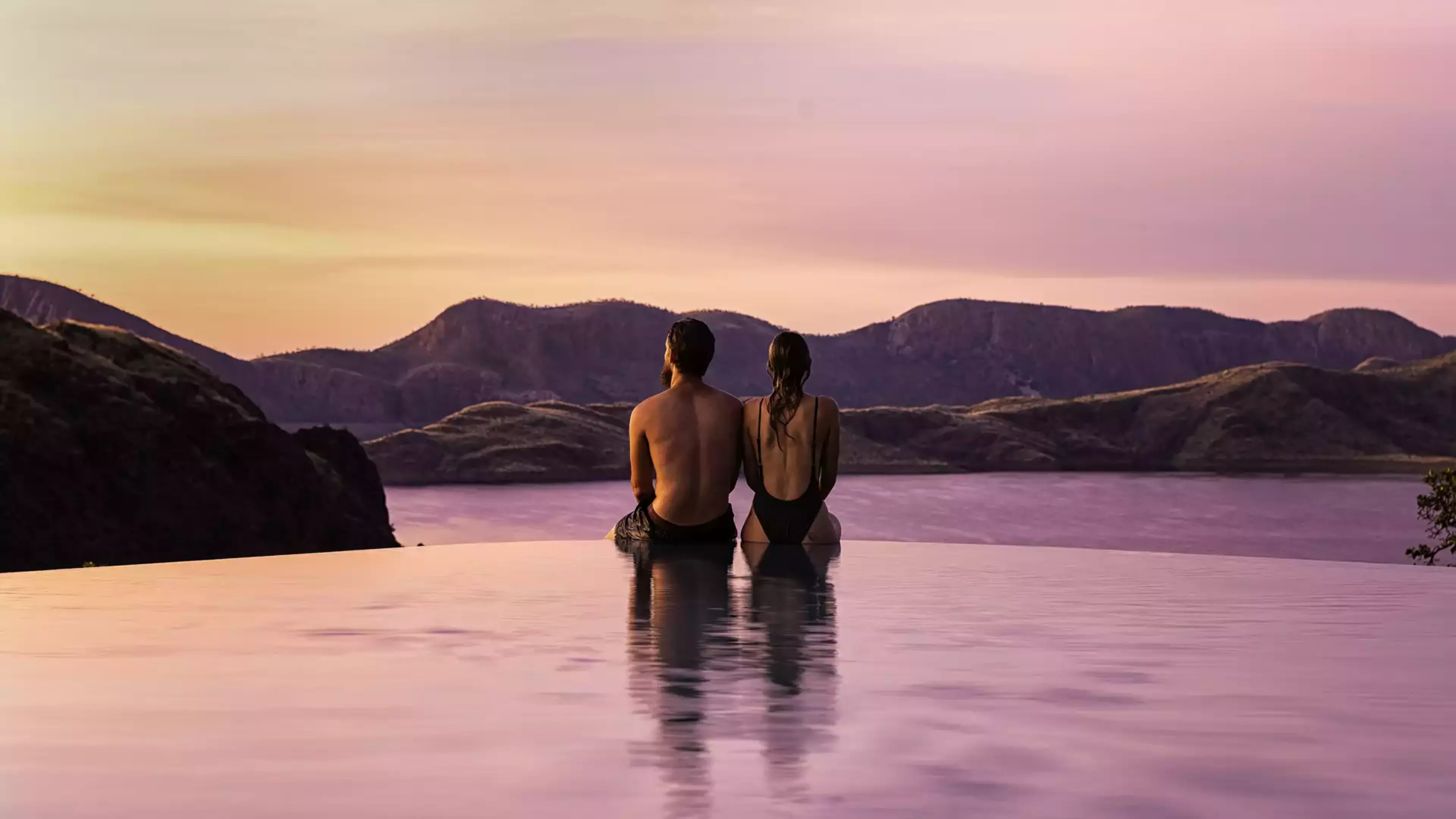 A couple relaxes at the edge of the famous infinity pool at Lake Argyle Resort, overlooking the vast, sunset-lit waters of the inland sea and the Carr Boyd Ranges.