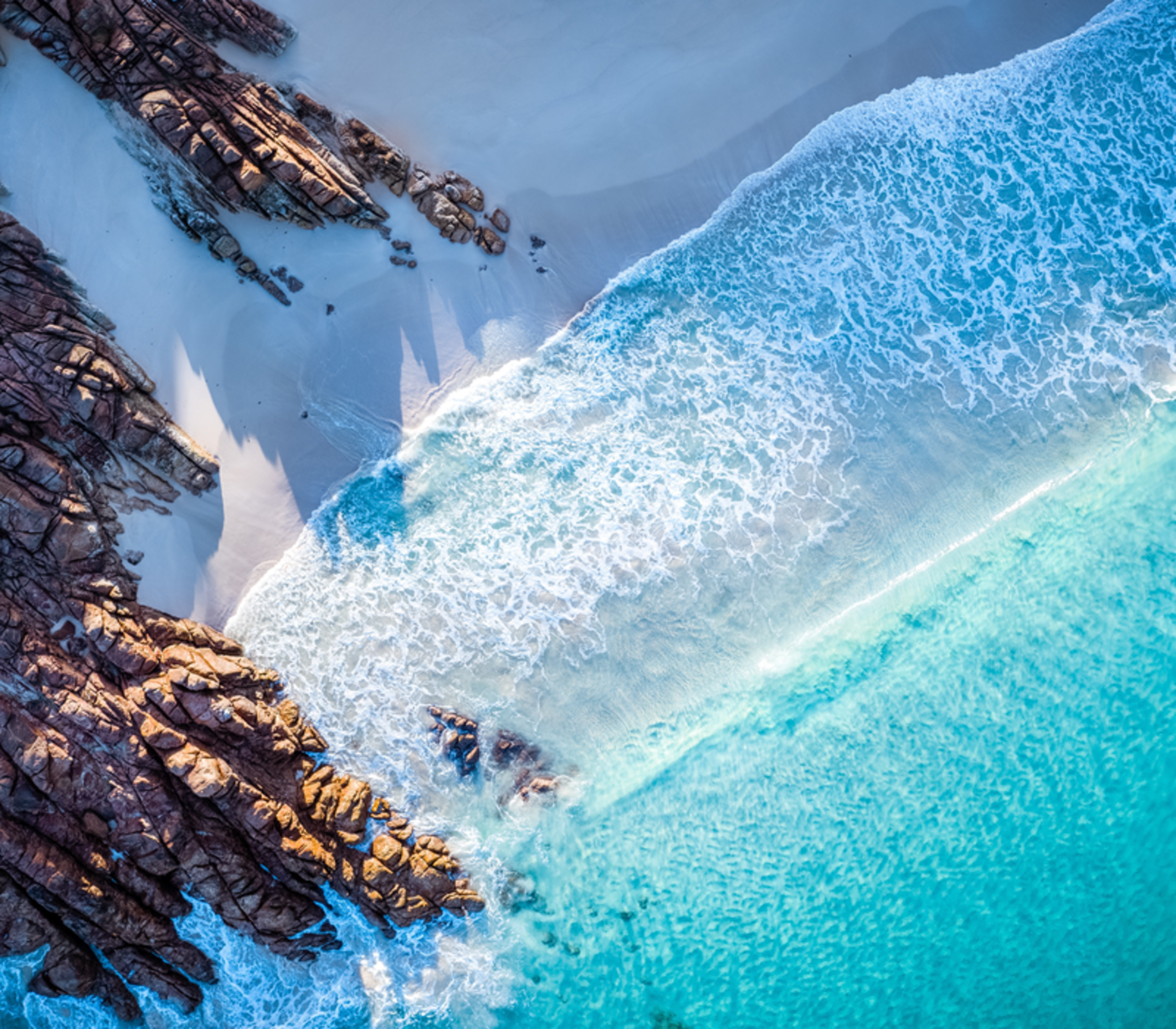 A dramatic top-down aerial view captures the white foam of the Indian Ocean crashing against the ancient red-tinted granite of Wyadup Rocks in Yallingup.
