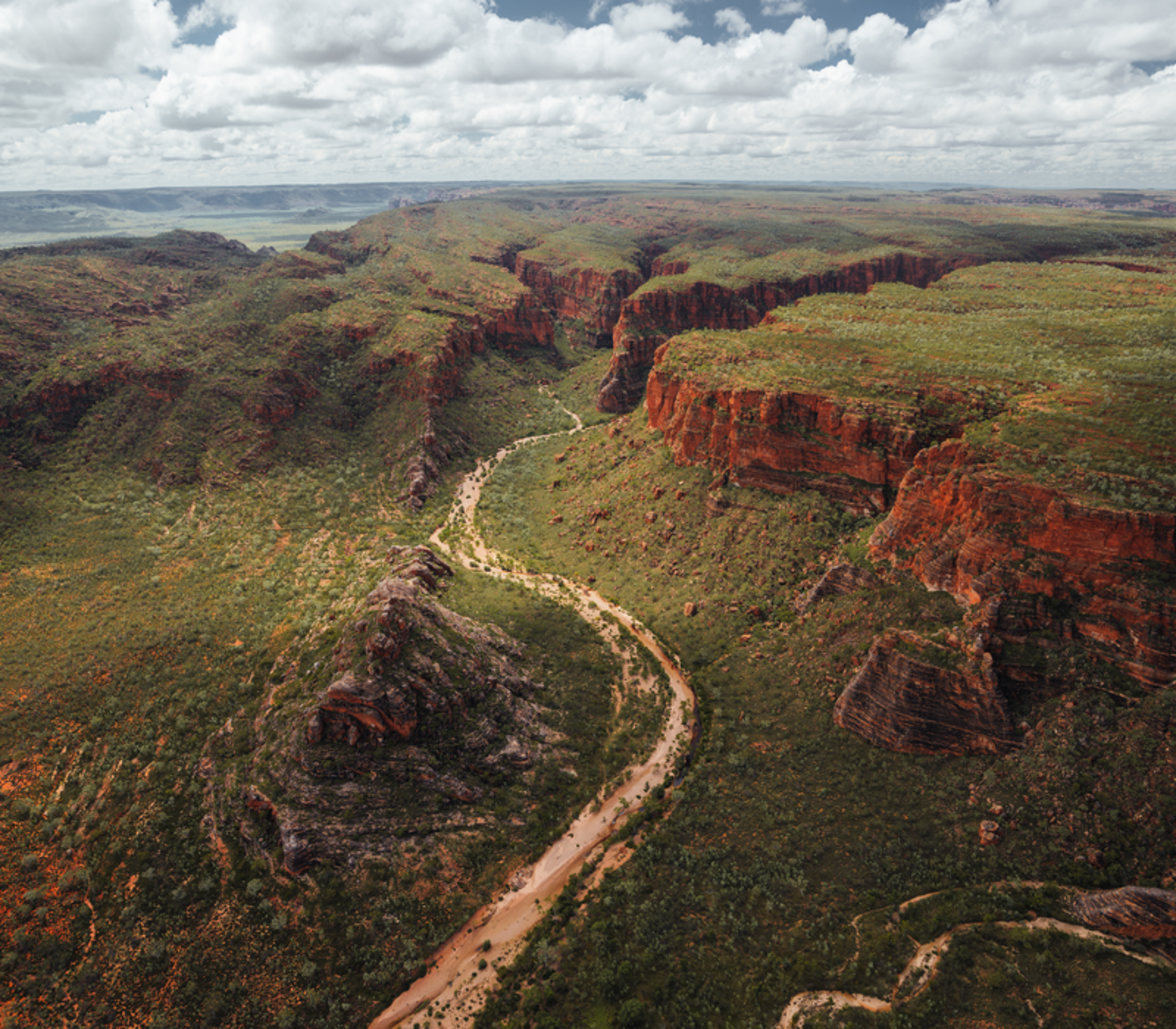 : Looking up from the sandy floor of a massive natural rock amphitheatre with towering, layered orange and red sandstone walls and a small patch of blue sky above.