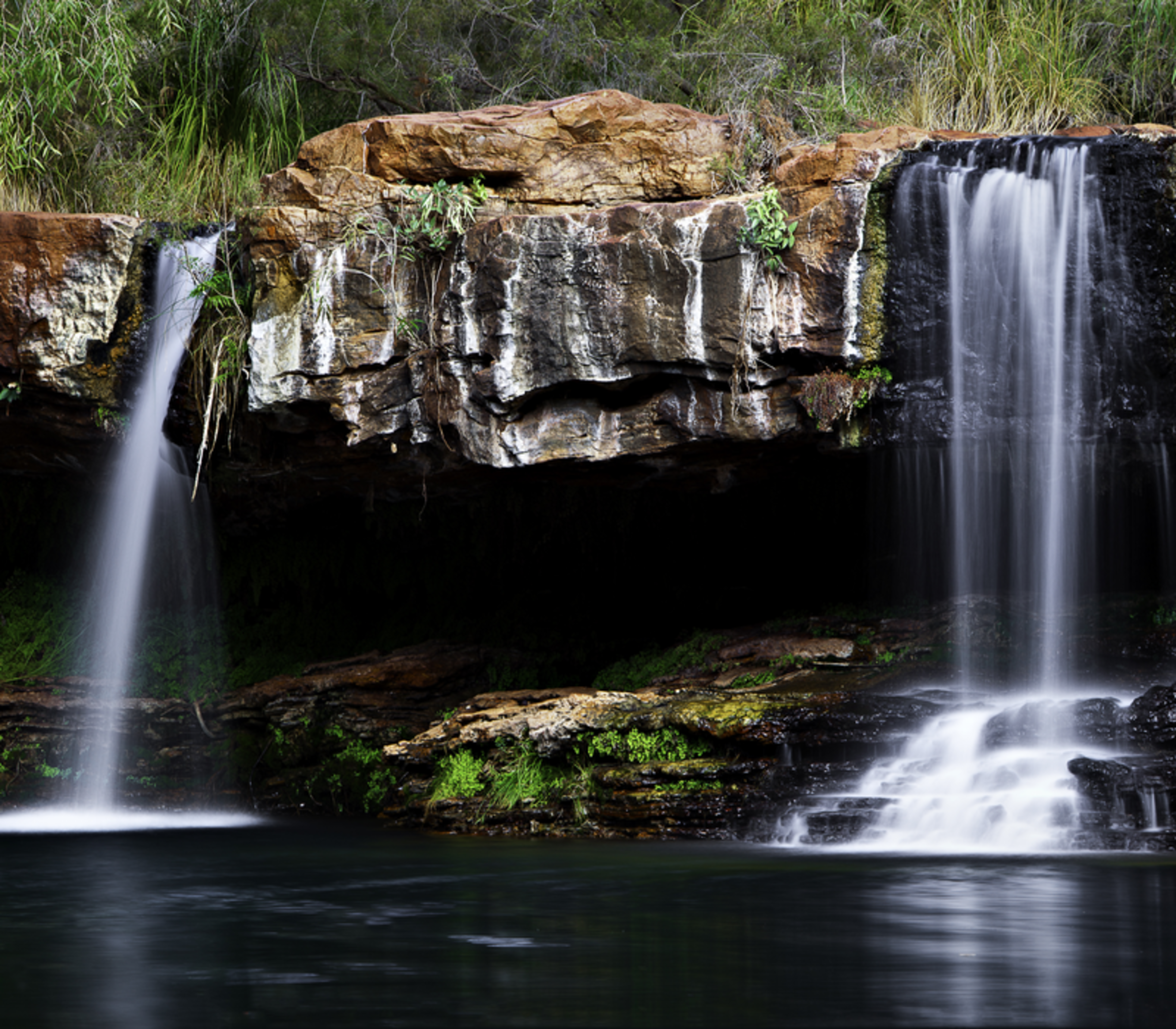 Twin waterfalls flowing over ancient red rocks into a dark green natural swimming pool surrounded by lush vegetation.