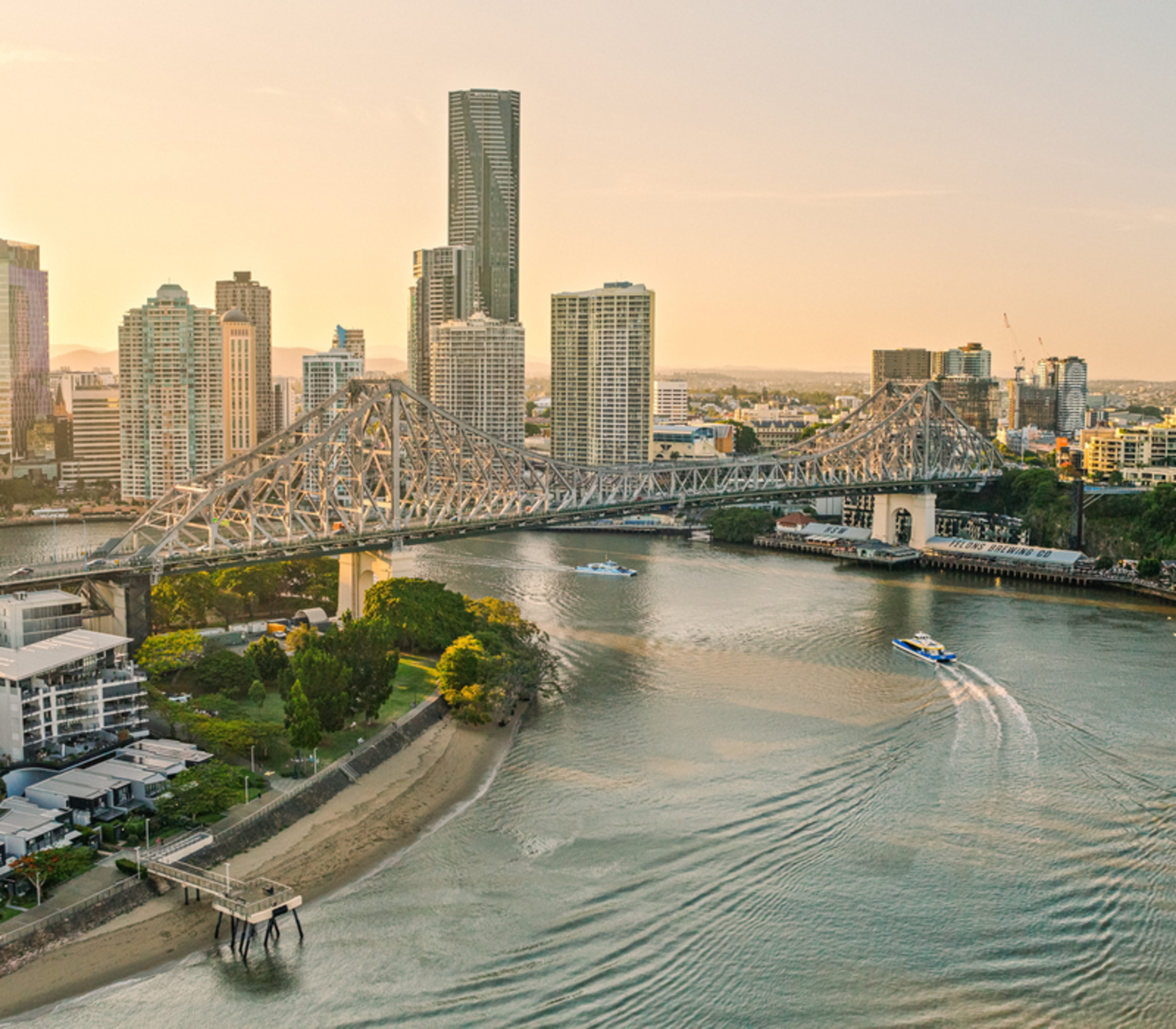 An aerial view of a wide river with several boats leaving white wakes, passing under a large steel bridge toward a city of tall buildings under a hazy yellow sky.