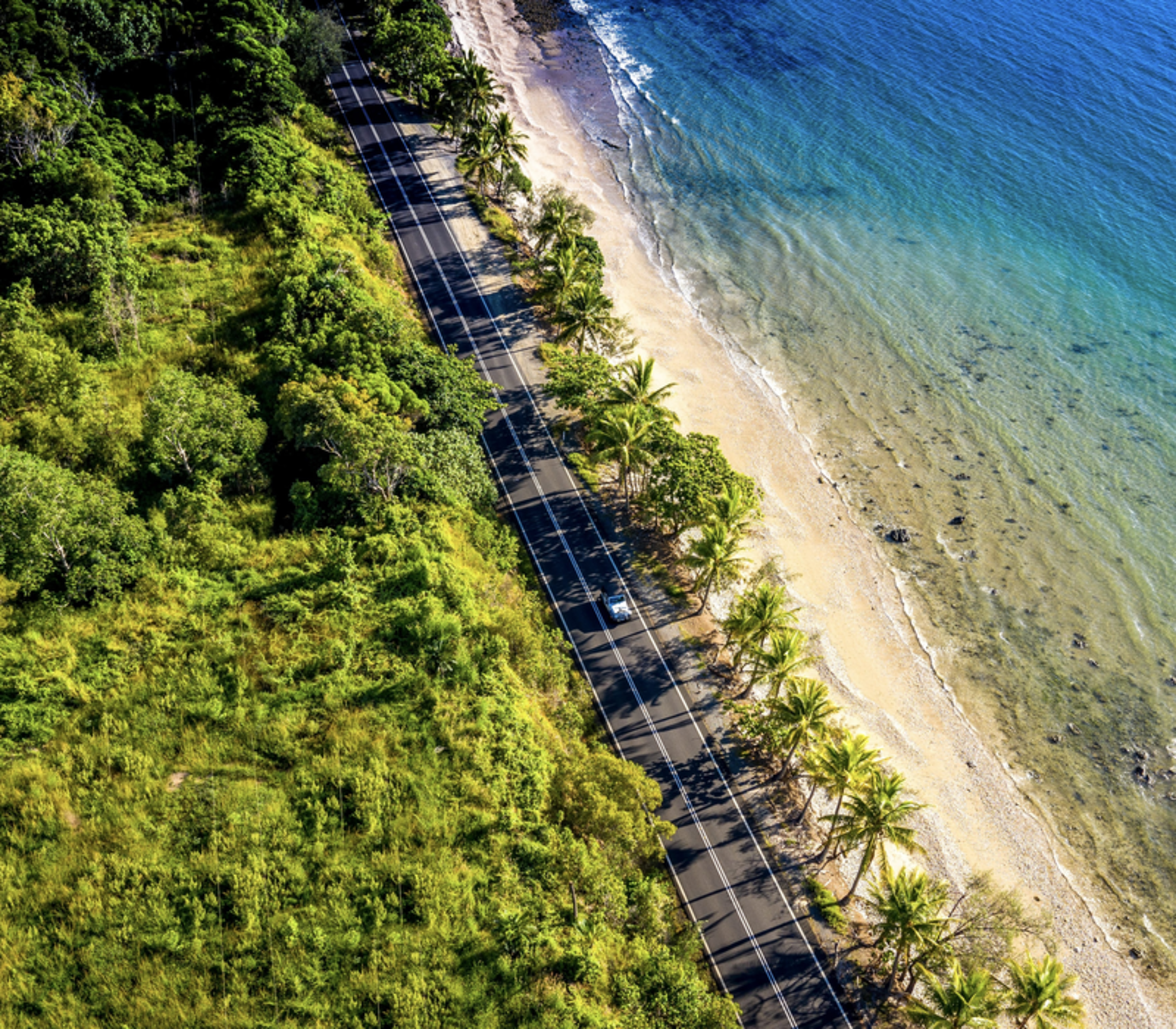 A high-altitude aerial view captures a white car traveling along a narrow asphalt road sandwiched between dense green tropical forest and a golden beach with turquoise water near Port Douglas.