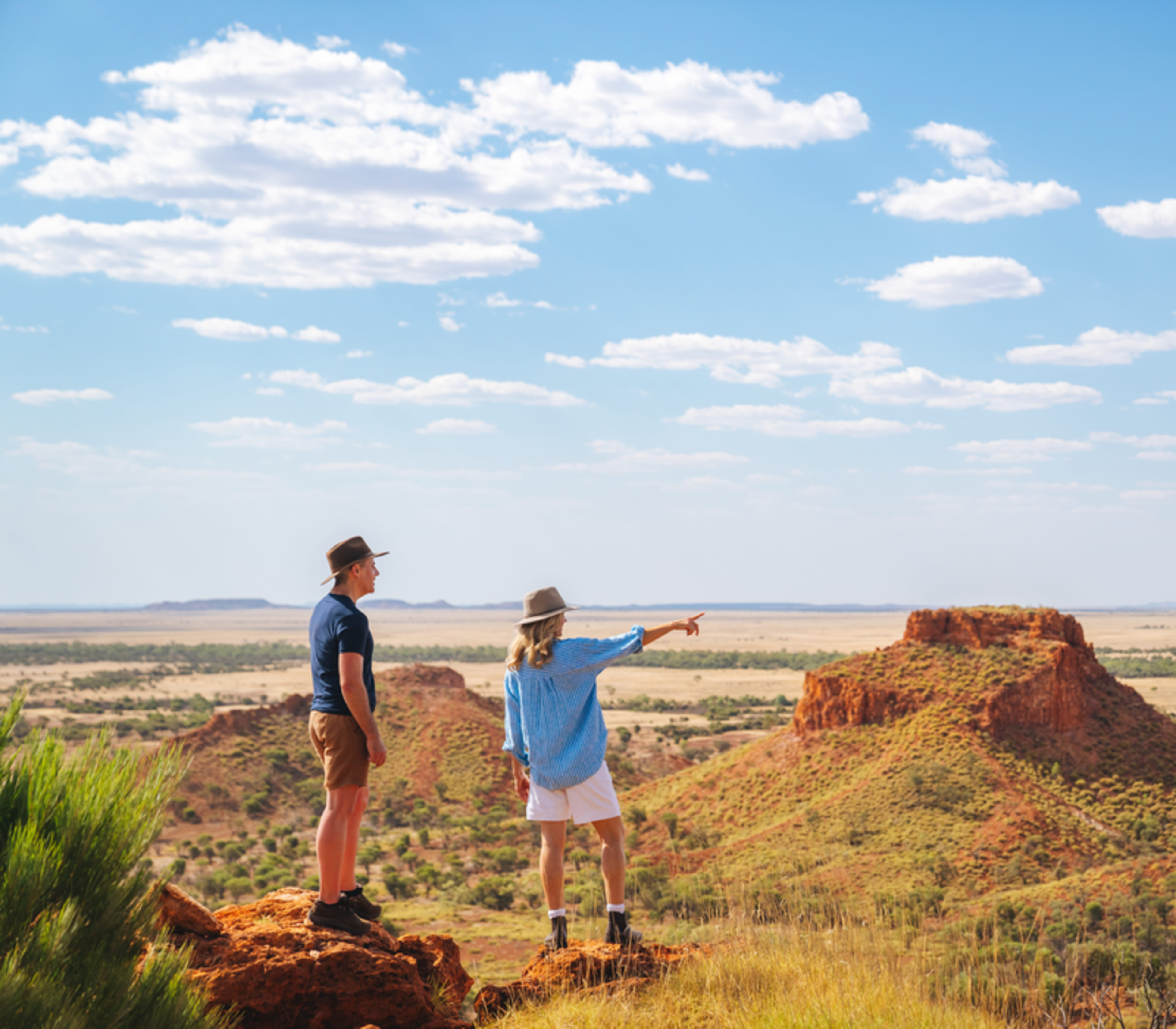 Two people standing on a rocky outcrop looking out over a vast outback landscape with red mesas.