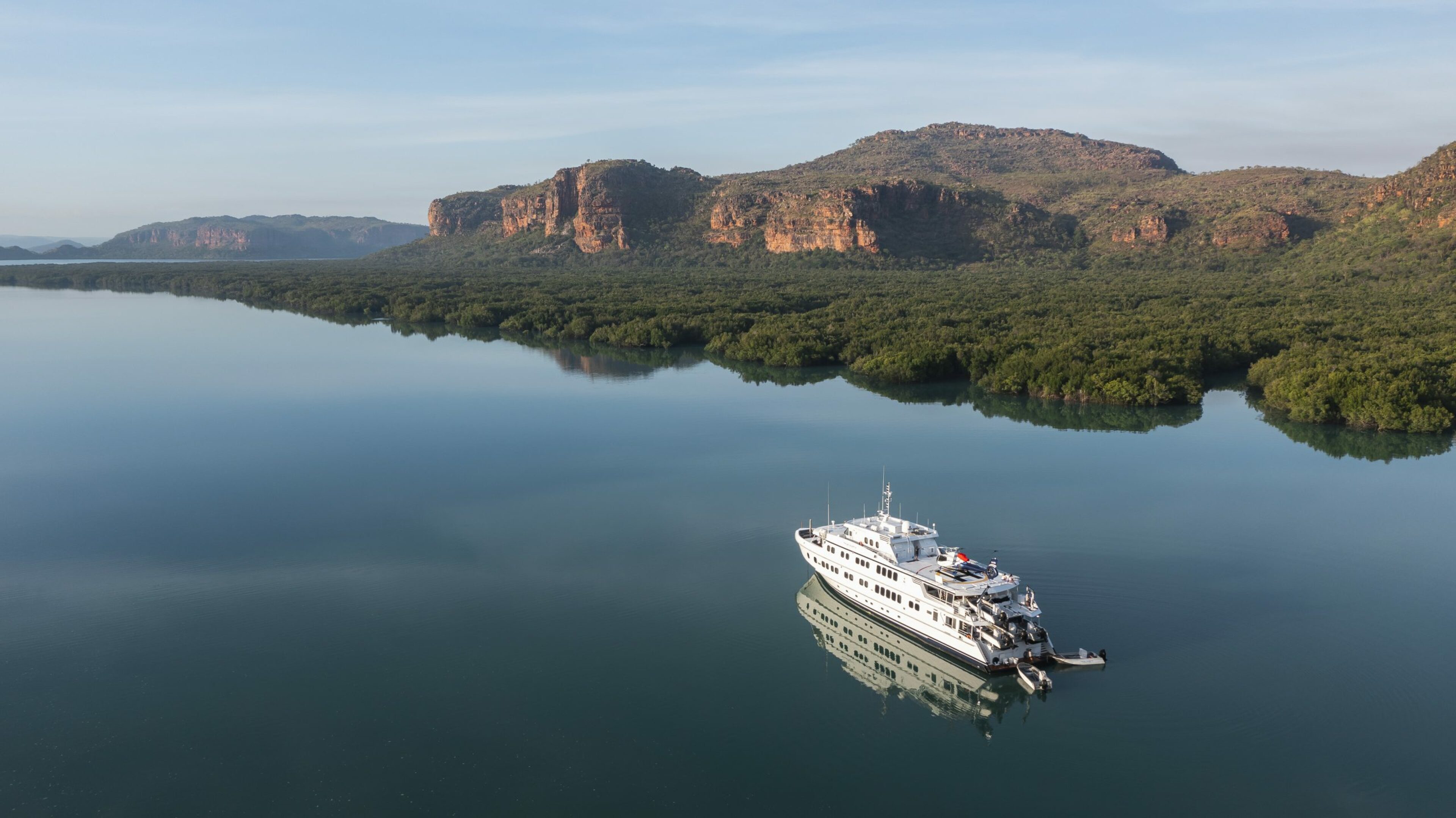 A True North cruise ship rests on perfectly calm water that reflects the sky next to a dense green mangrove forest and hills in the Kimberleys.