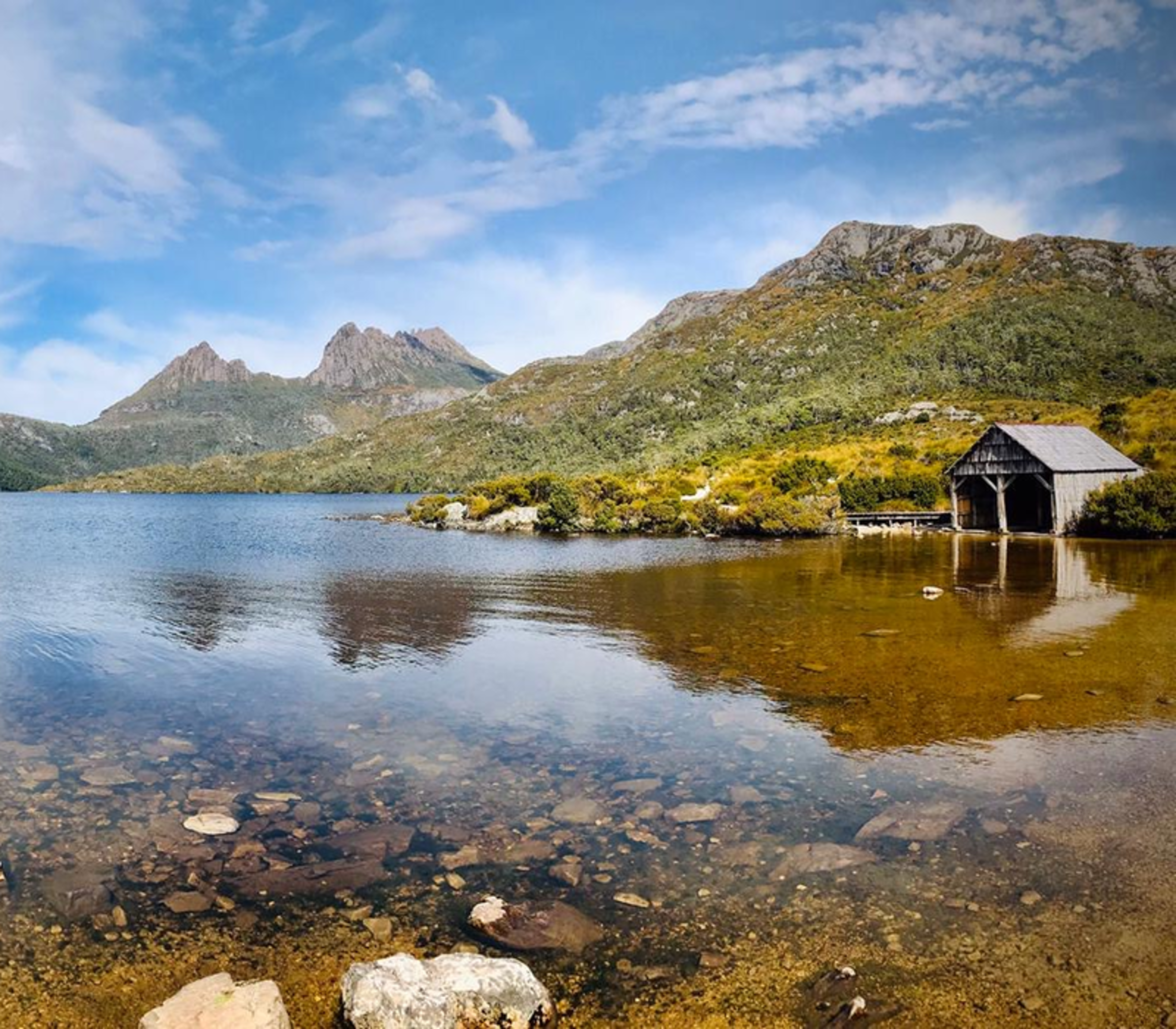 A wide landscape view of Dove Lake in Tasmania featuring the historic wooden boatshed and Cradle Mountain under a blue sky.