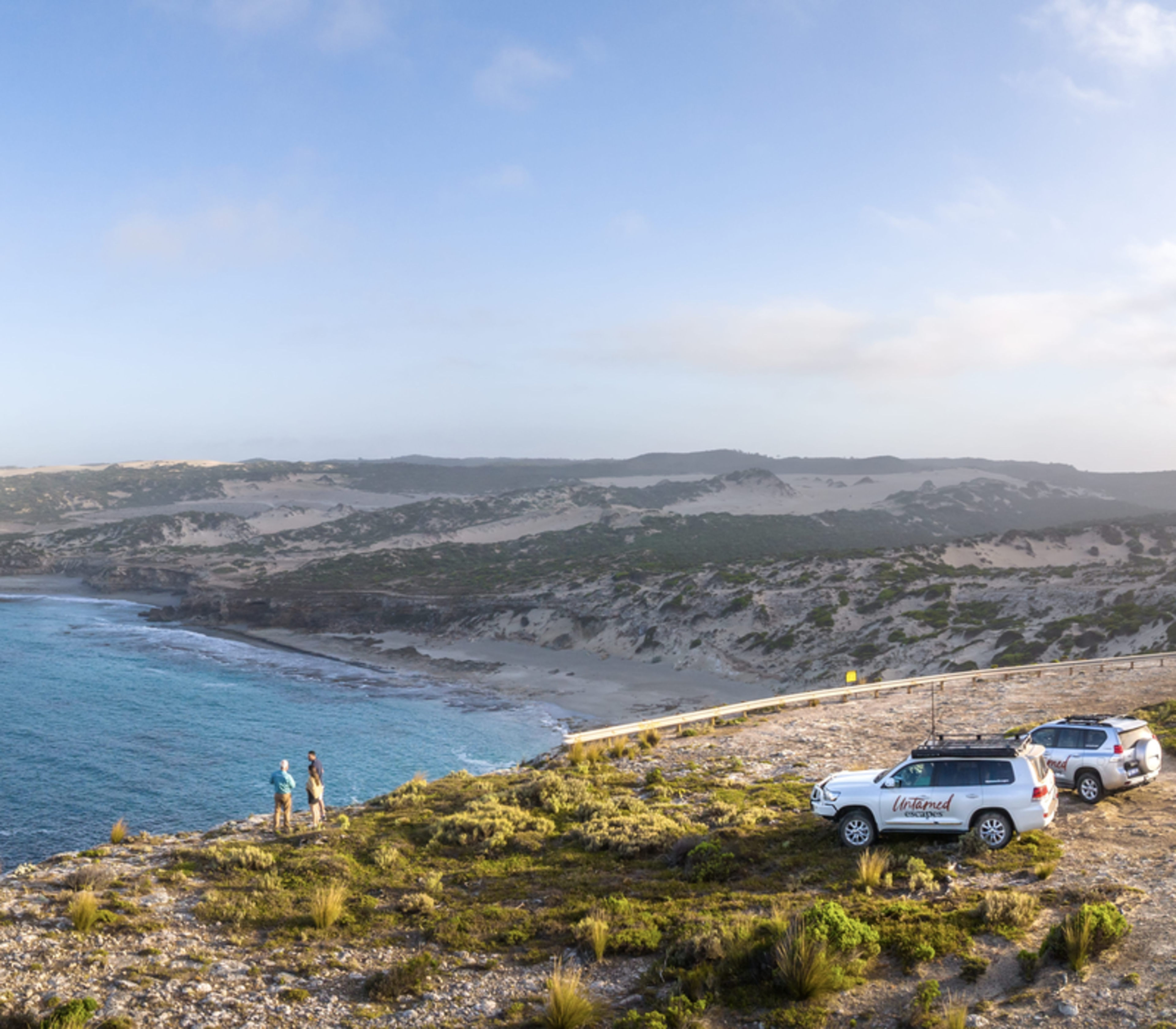 Two 4WD vehicles parked on a high cliff overlooking a rugged bay with turquoise water and sandy beaches.