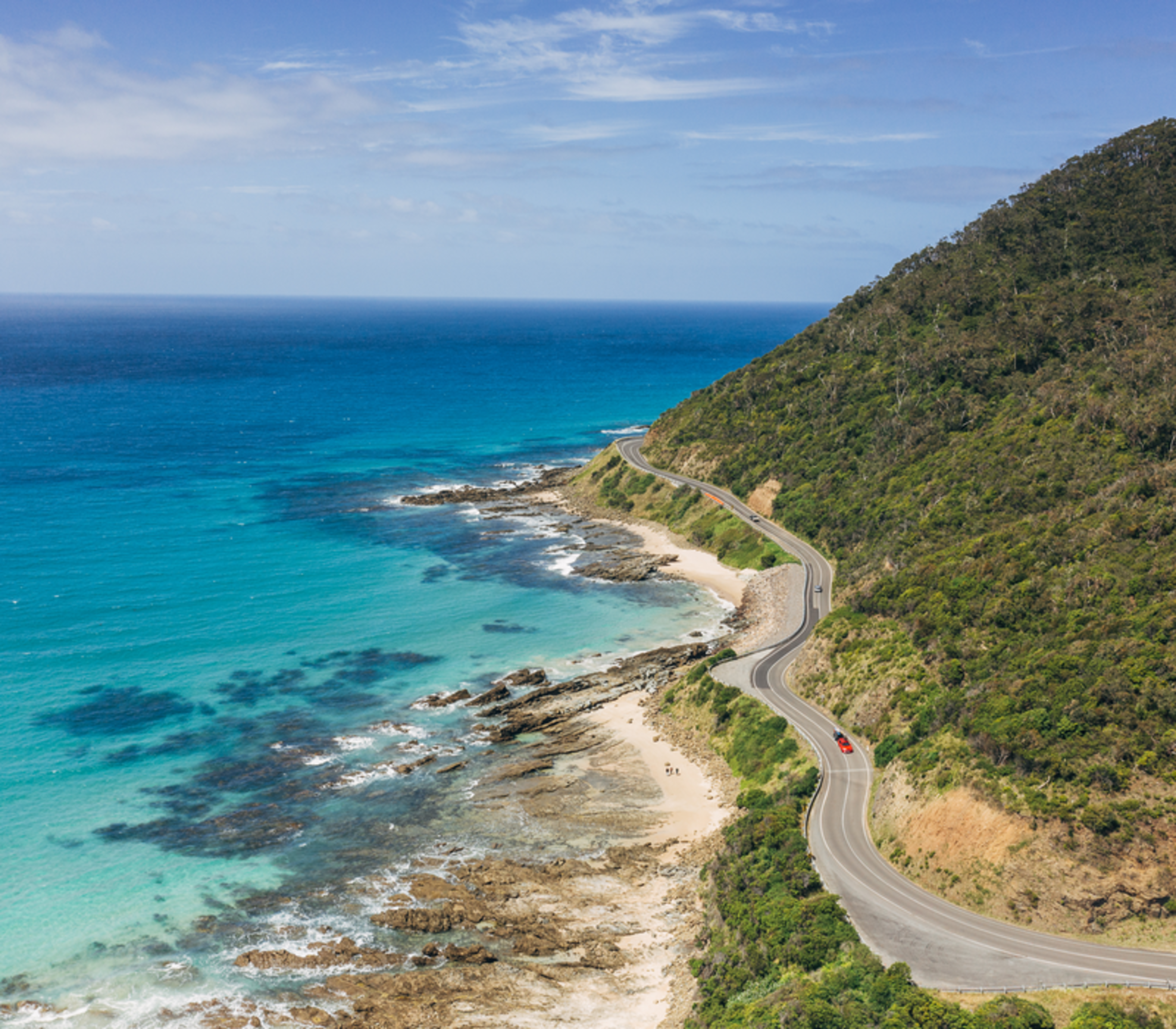 High-angle view of the Great Ocean Road winding along a turquoise coastline and green hills.