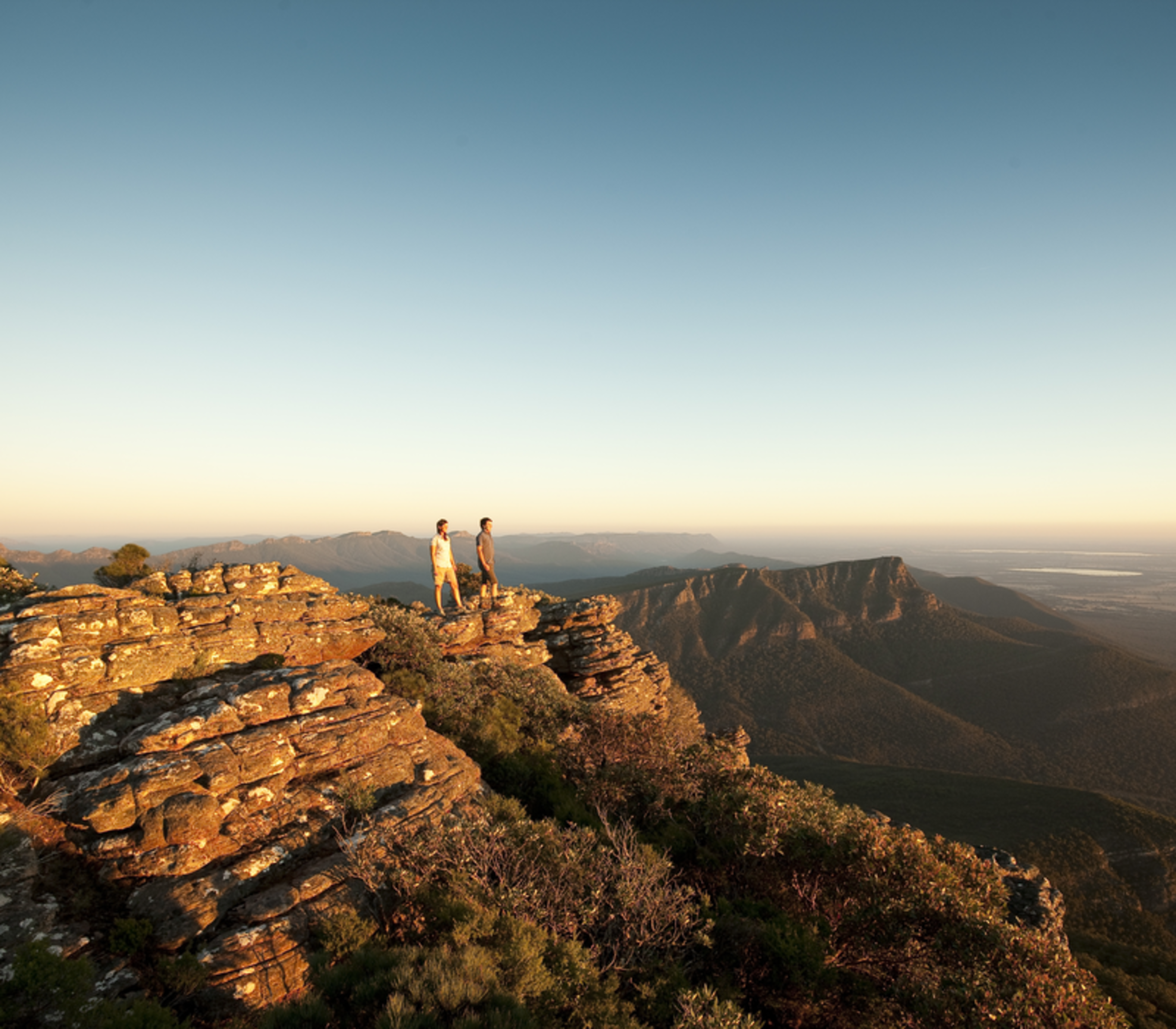 Two people stand atop a flat, weathered sandstone outcrop overlooking a vast, rolling forested valley and distant mountain ranges bathed in the soft golden light of a setting sun in The Grampians.