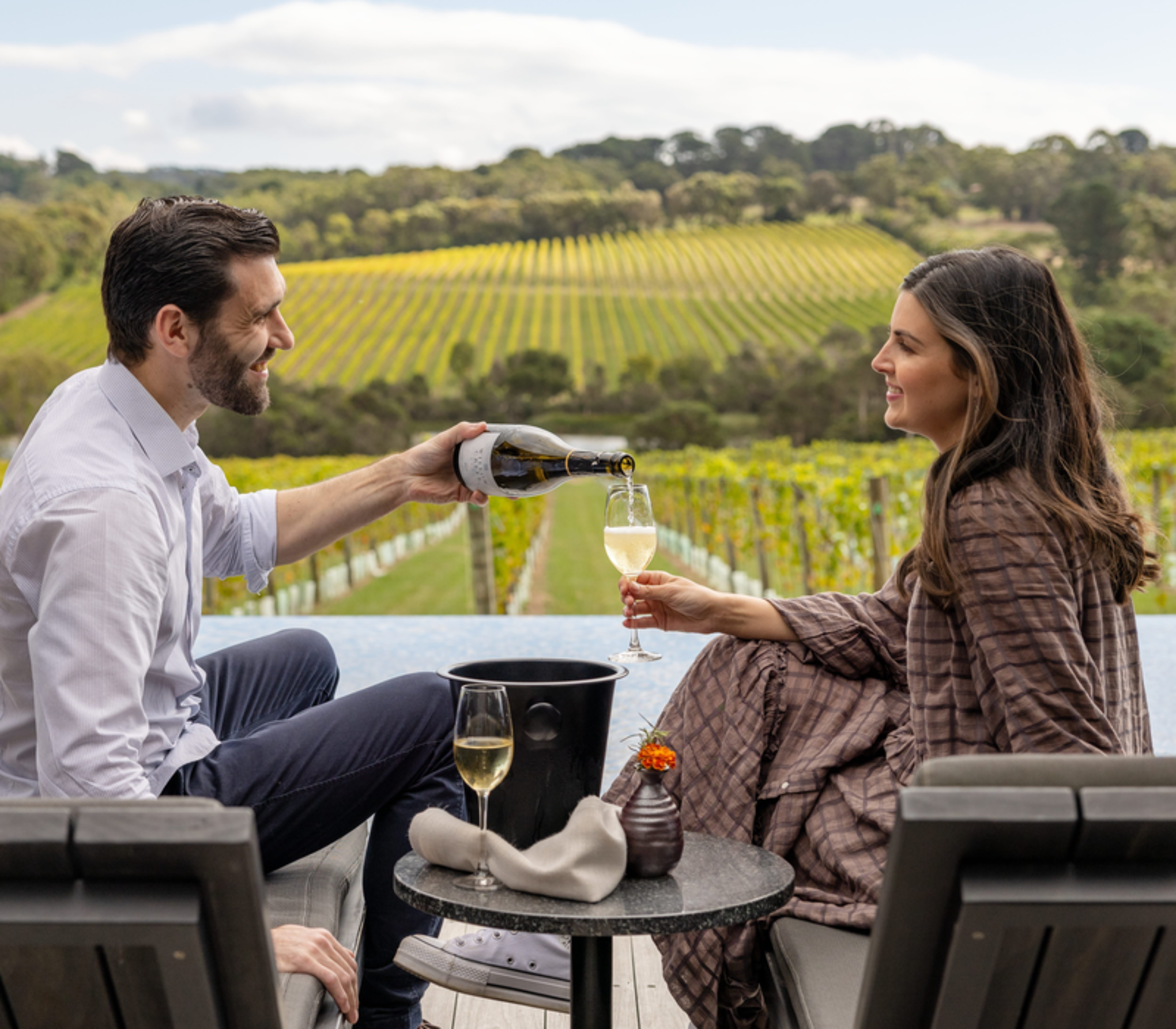 A man pouring wine for a woman sitting on lounge chairs next to an infinity pool overlooking a vineyard.