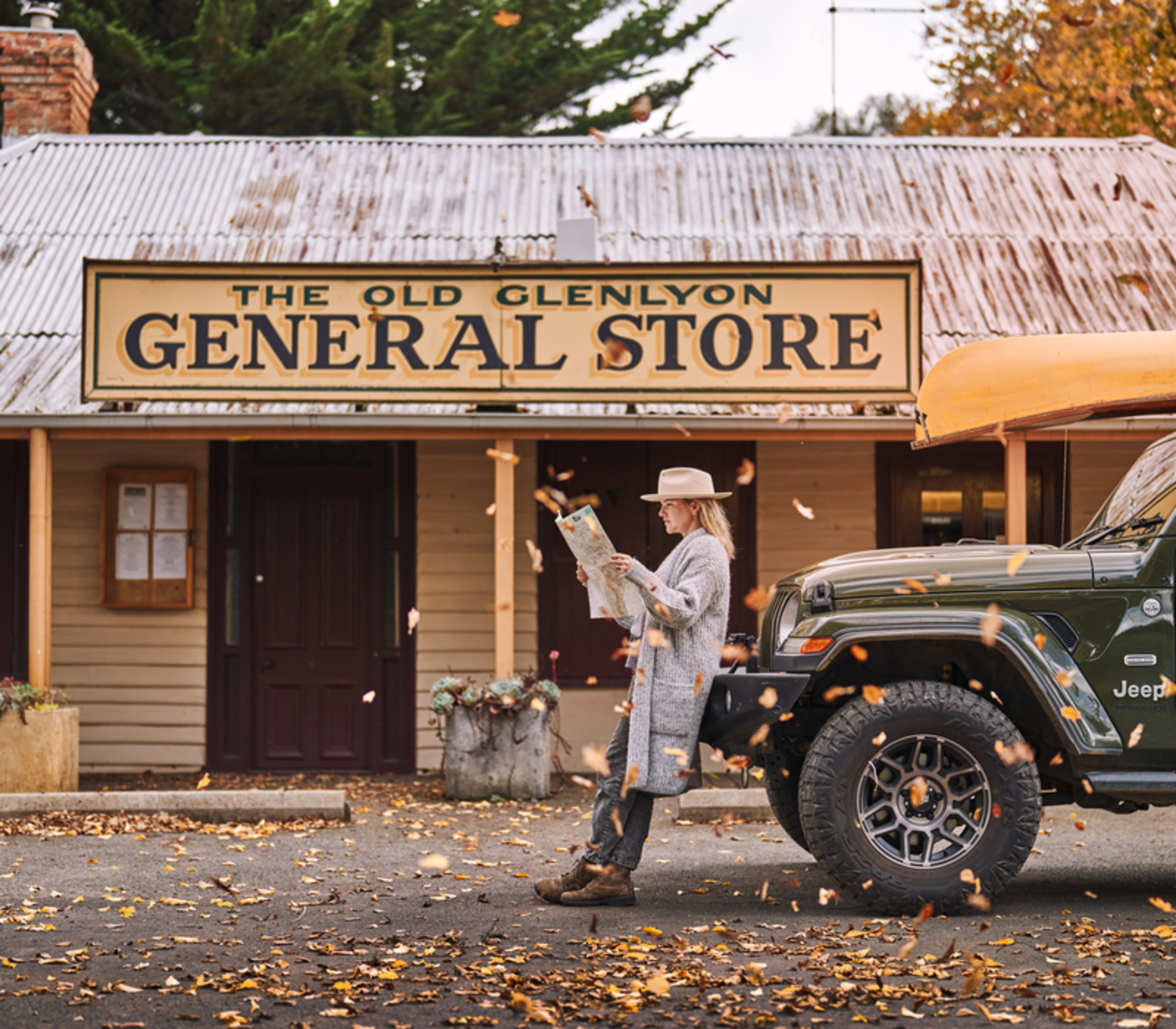 A woman in a hat leans against a 4WD with a canoe, reading a map outside a rustic general store.