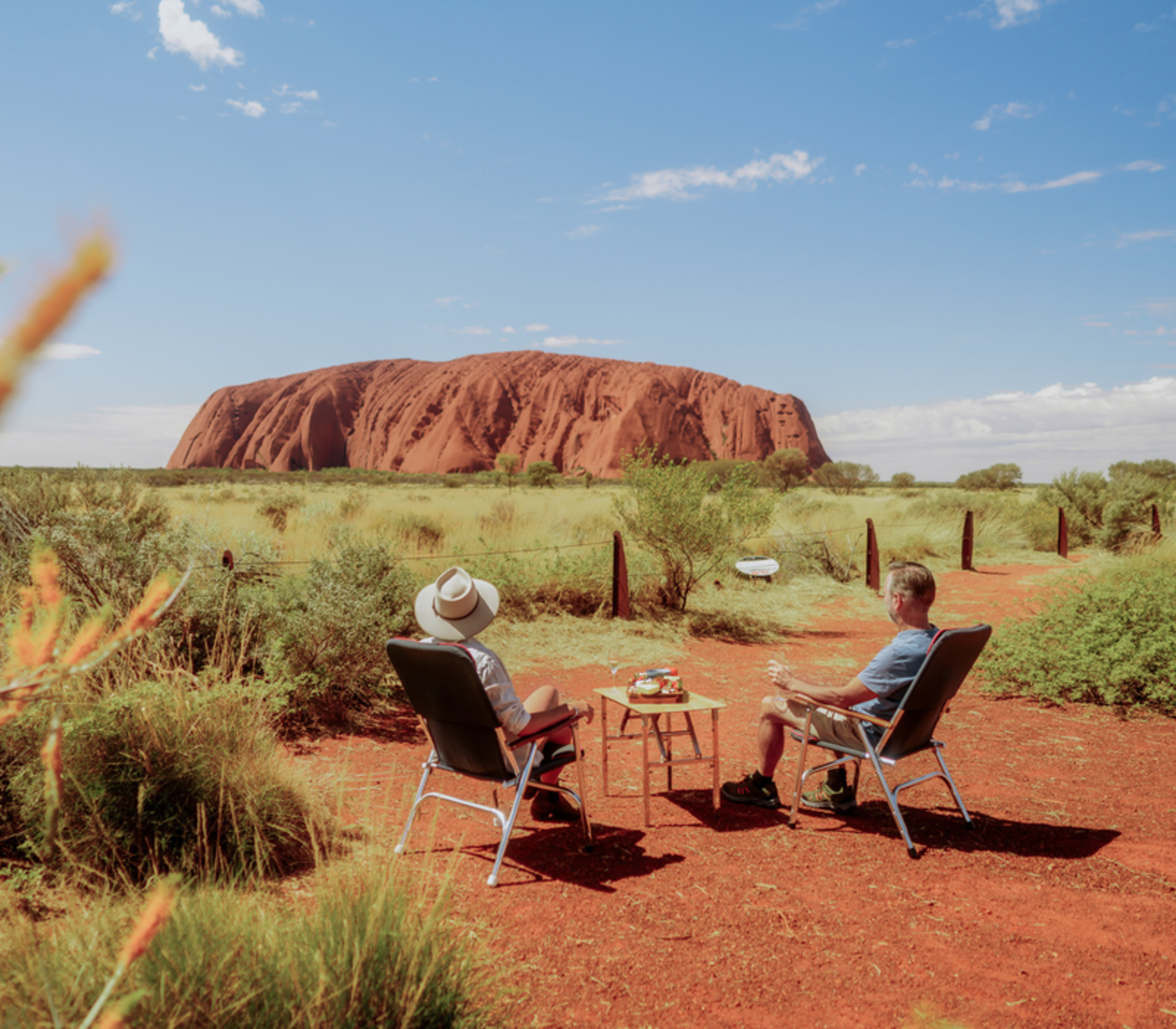 Two people sitting in camping chairs with a small table of food overlooking Uluru in the desert.
