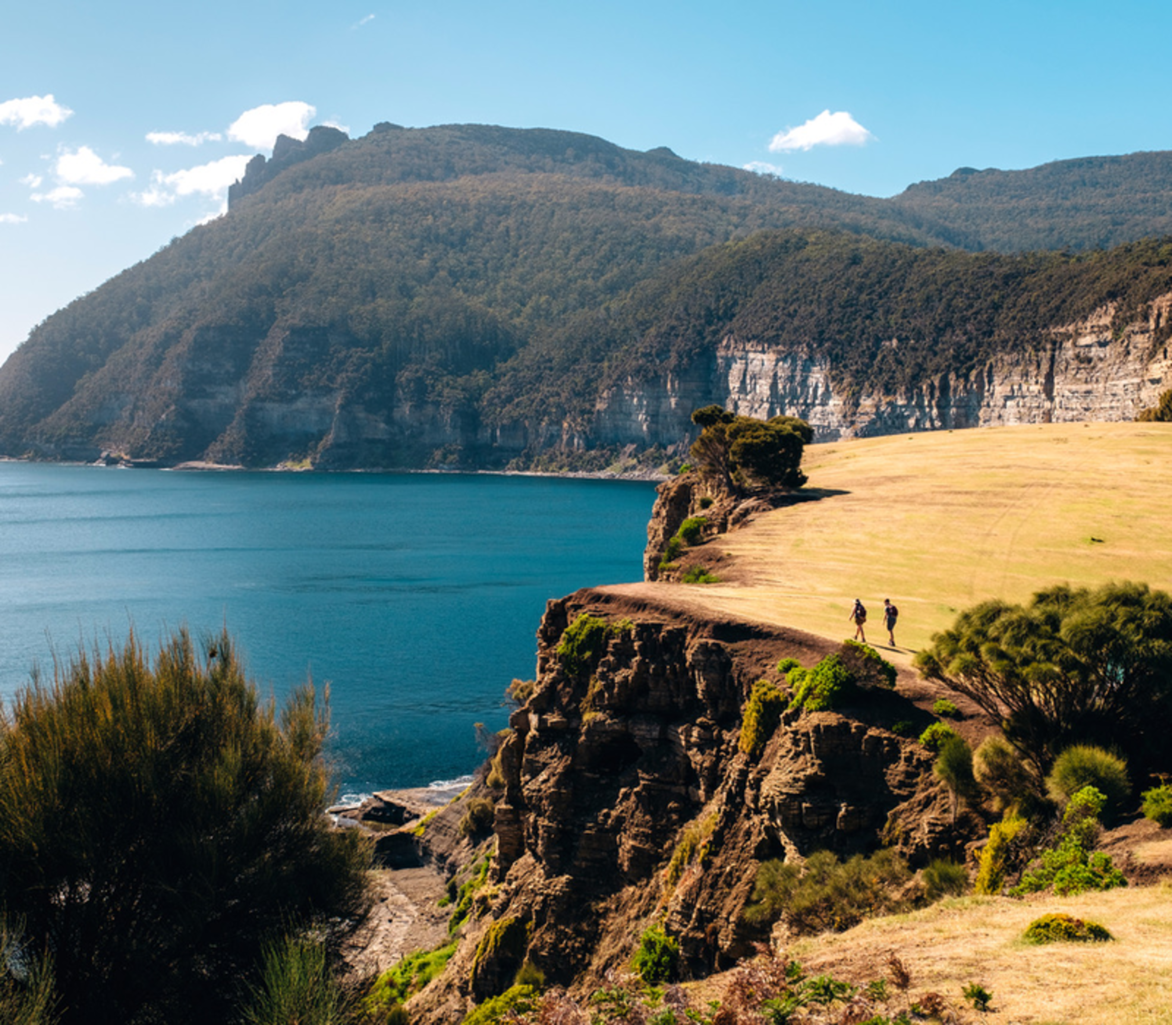 Two hikers walking on a grassy cliff edge overlooking a deep blue ocean bay.