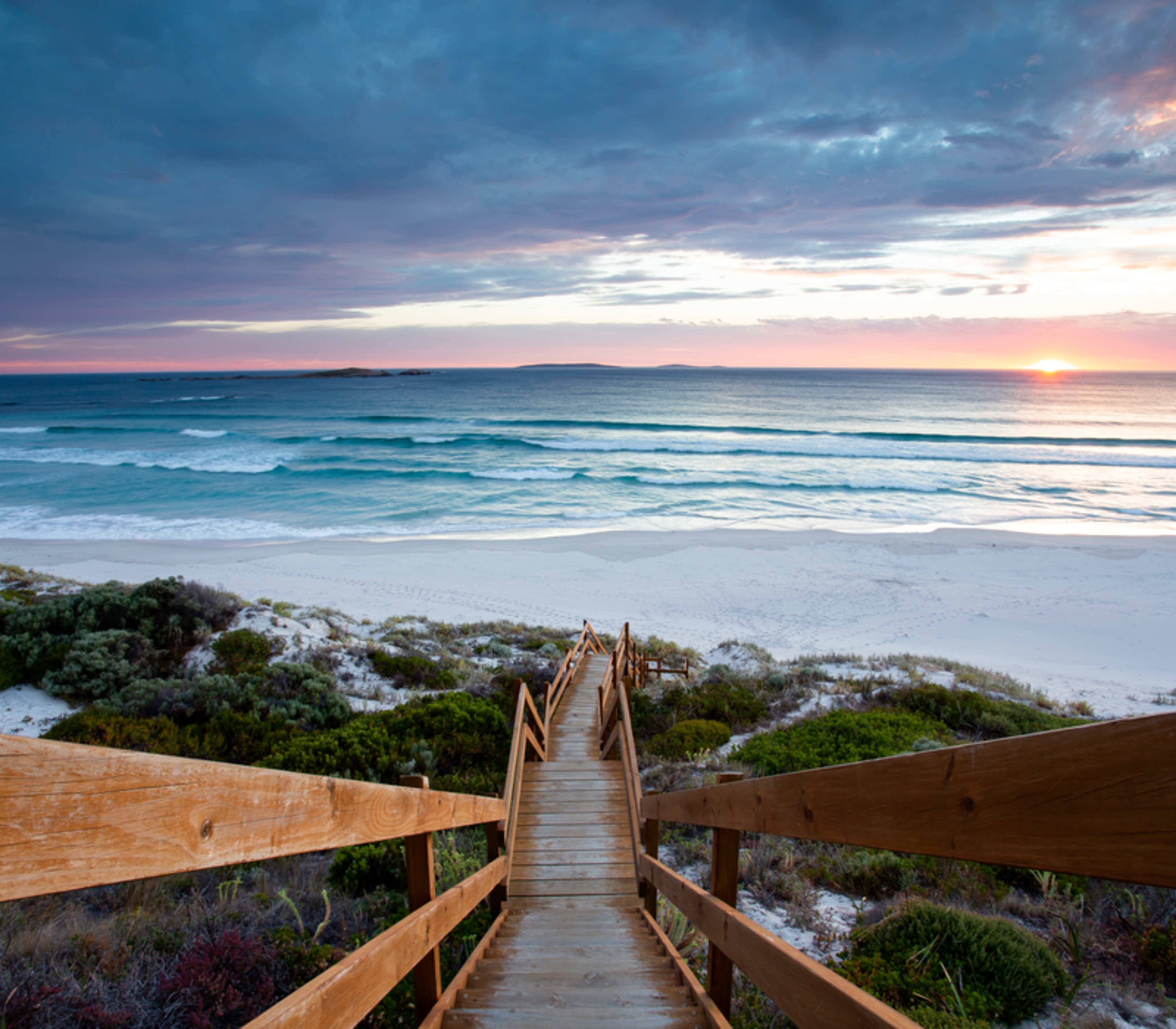 A wooden staircase leading down a scrub-covered dune to a wide white beach and ocean.