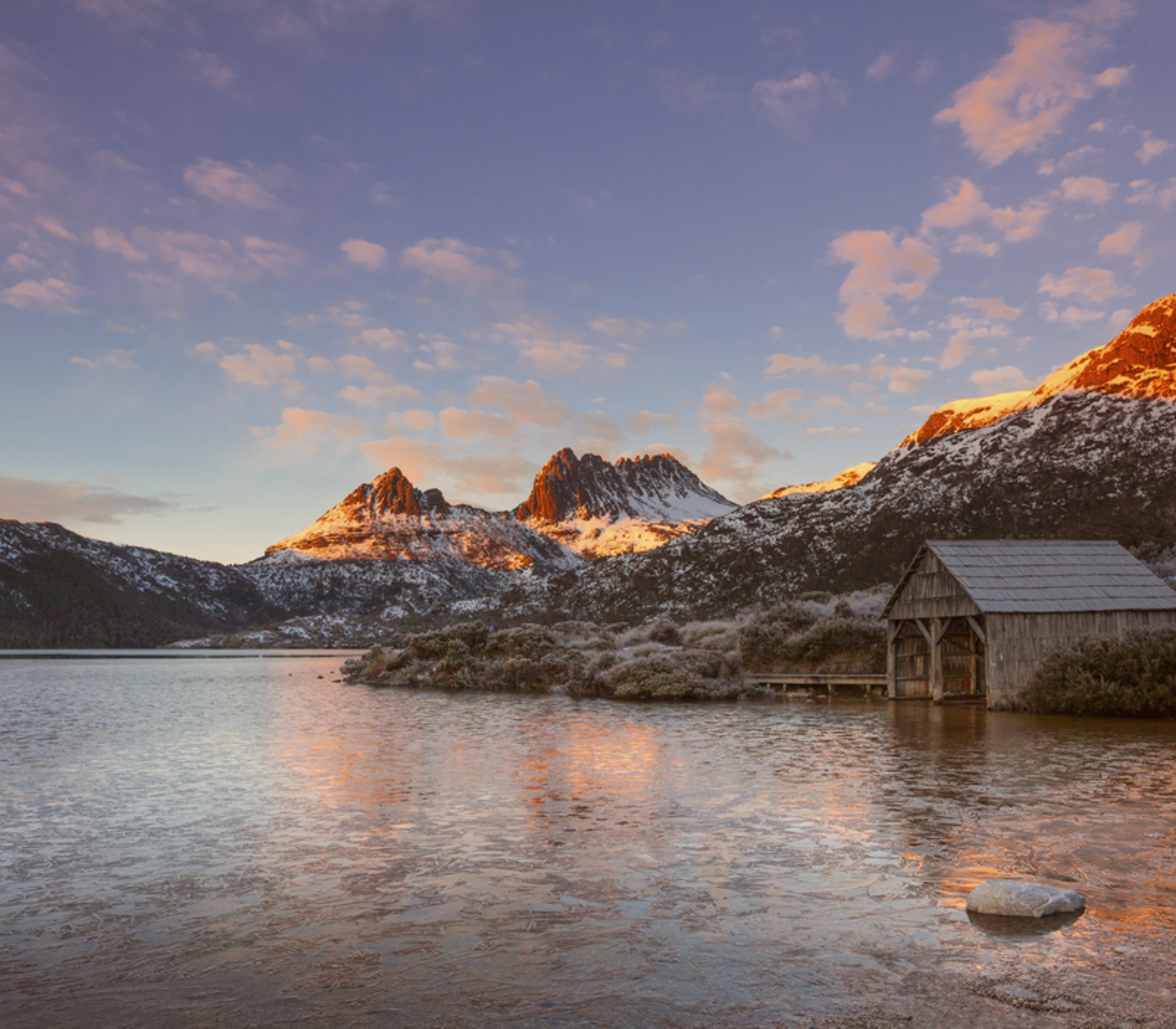 The iconic wooden boat shed on the shore of Dove Lake with Cradle Mountain reflecting the colors of a winter sunset.