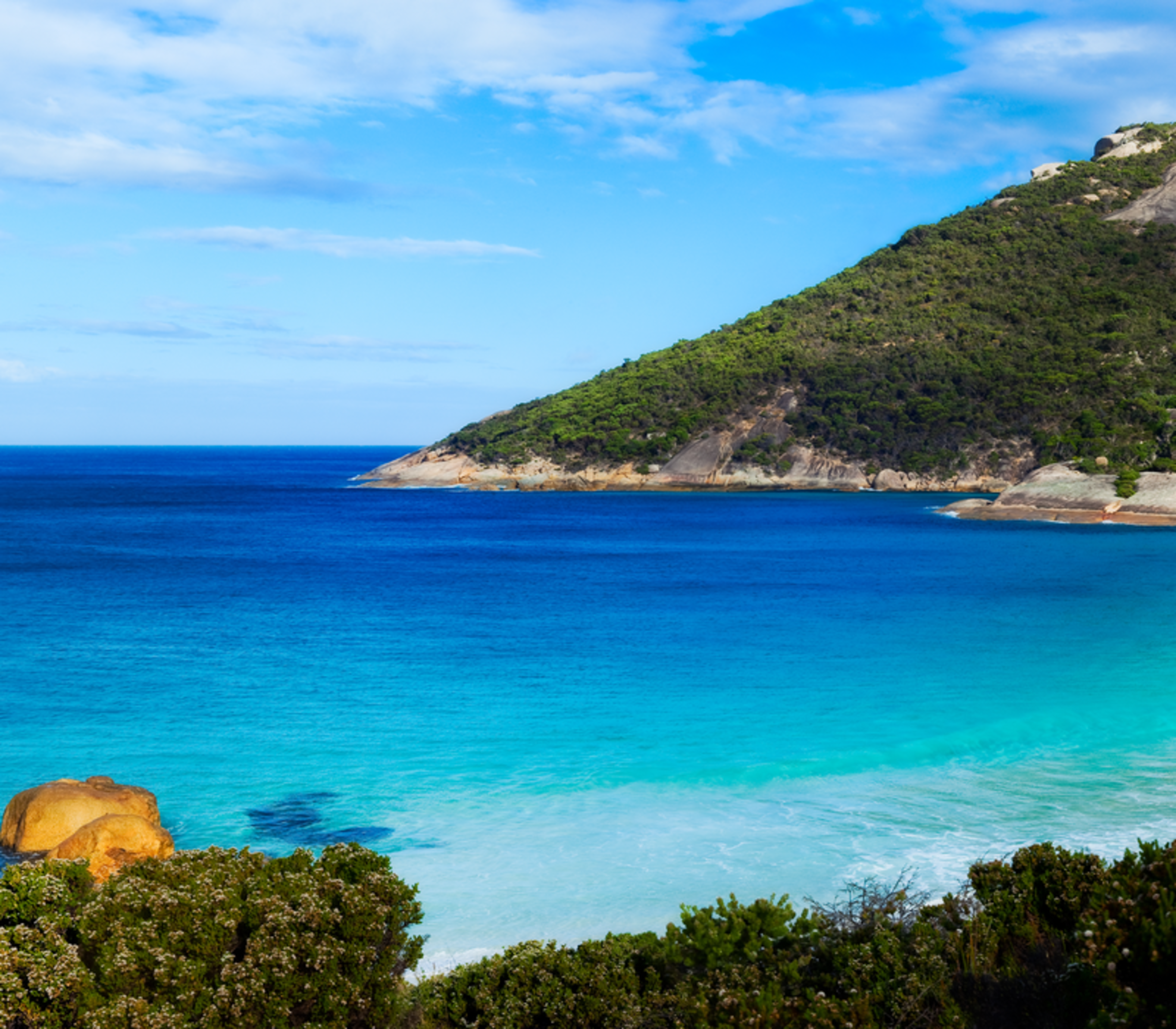 Wide panoramic view of a pristine white sand cove with turquoise ocean water and green coastal hills under a blue sky in Australia.