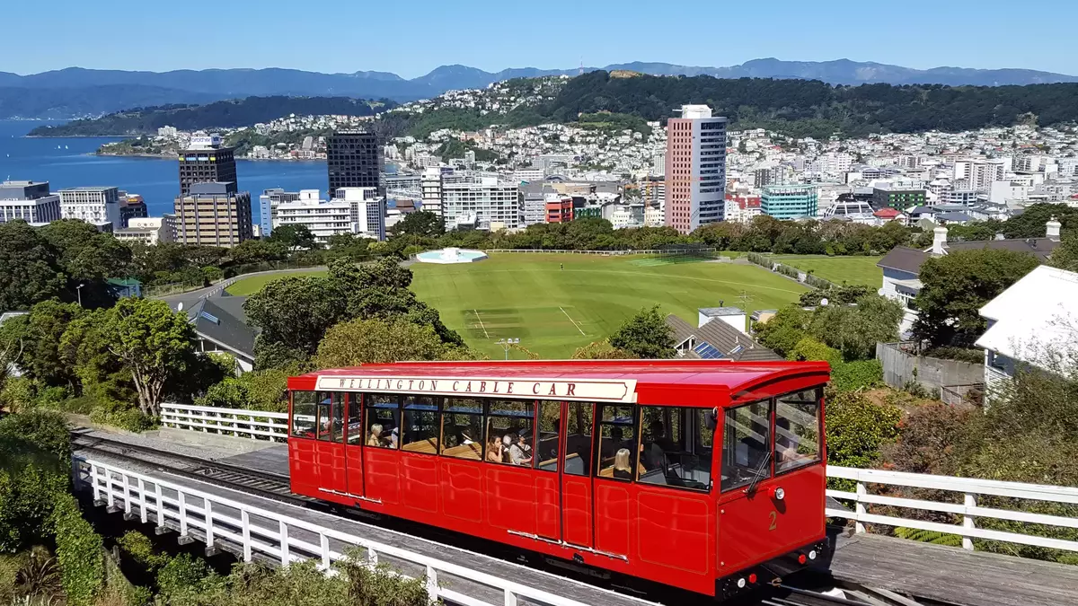 The bright red Wellington Cable Car travels down its tracks overlooking Wellington city, the harbor, and the surrounding hills under a clear blue sky. Main Image