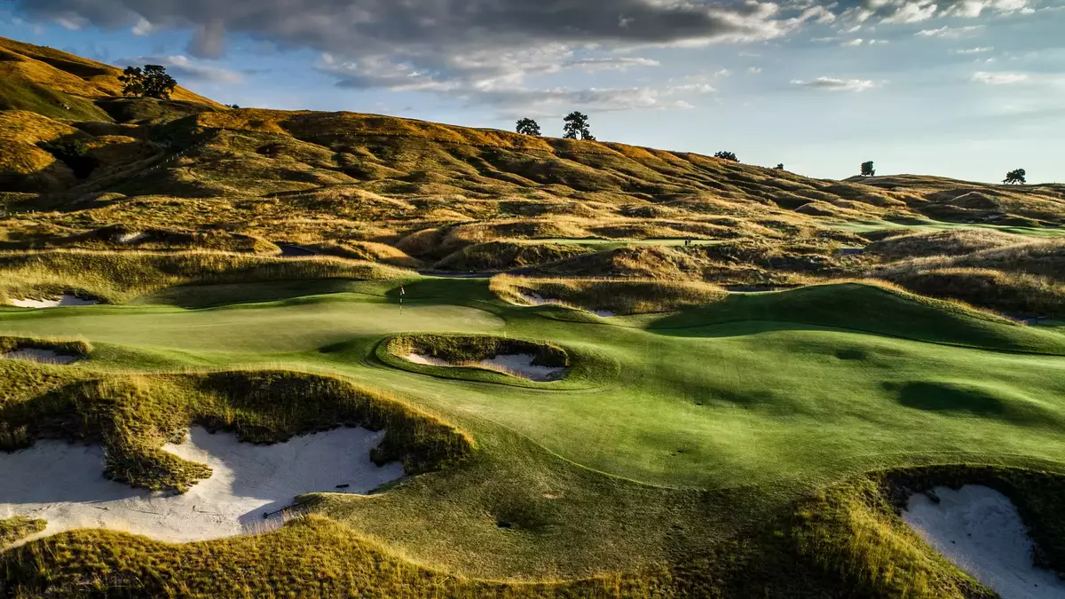 Golf course fairway with green, sand bunkers, and hills under cloudy sunset sky. Main Image