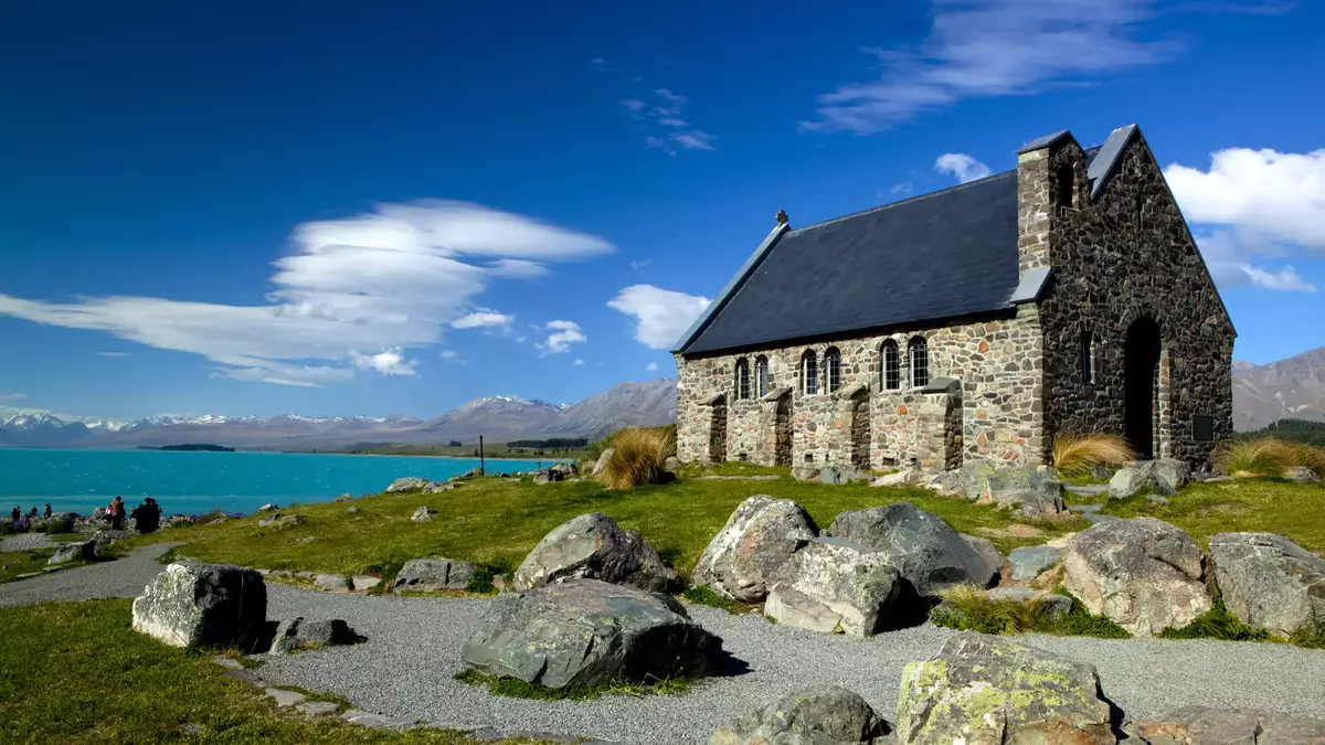 Stone church near turquoise lake with mountains and blue sky in the background. Main Image