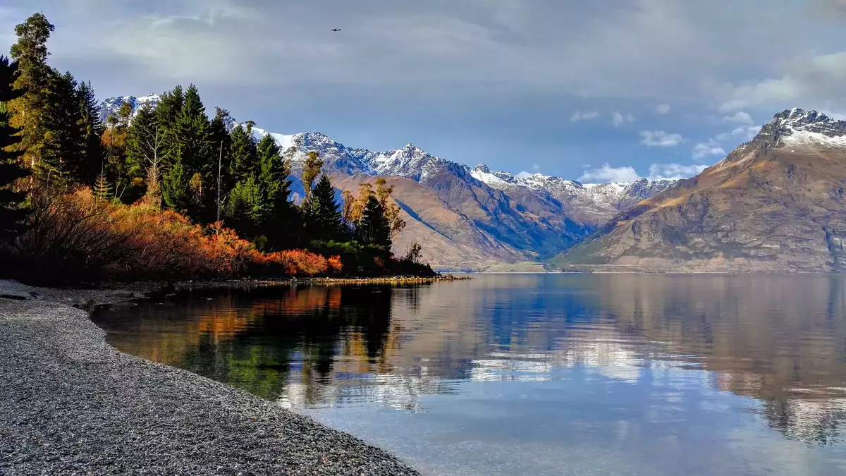 Lake with clear water reflecting snow-capped mountains and trees on a partly cloudy day. Main Image