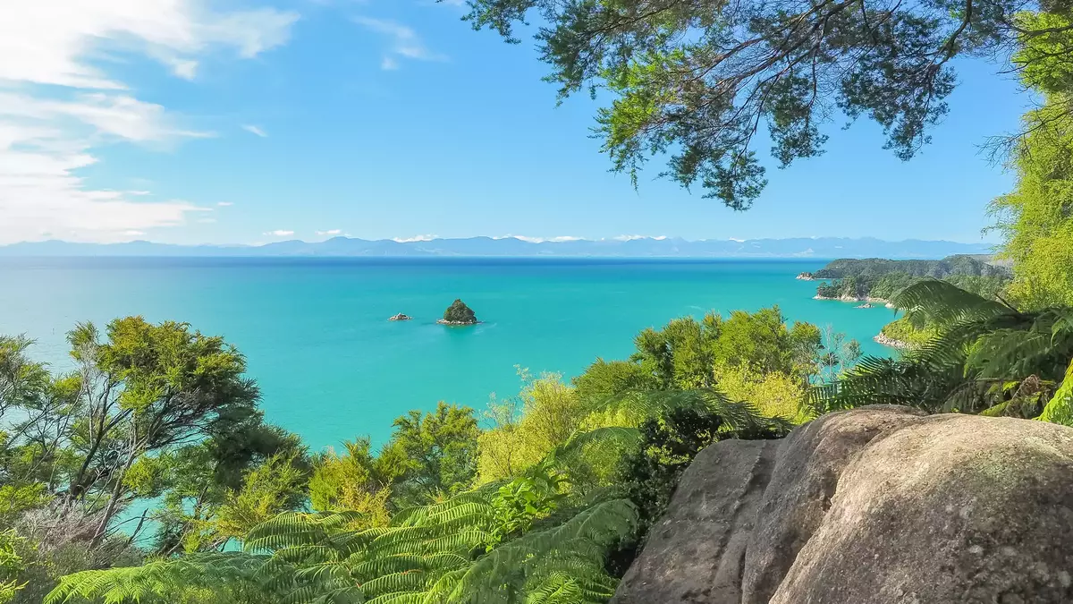 Looking through green trees at a small rocky island in a bright blue sea. Main Image