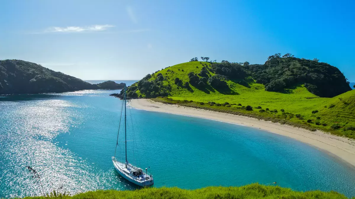 Bright blue sky with green verges, a curved sandy beach and the blue sea with a small boat in the centre  Main Image