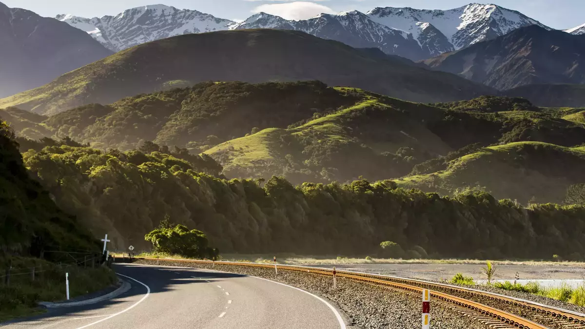 Road and railway track next to green hills with snowy mountains in the background. Main Image
