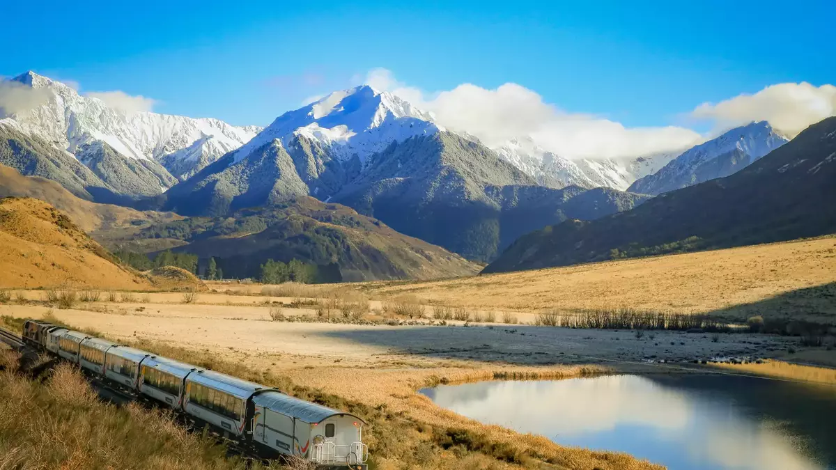 Train passing through valley with snow-covered mountains and lake reflecting sky and clouds Main Image